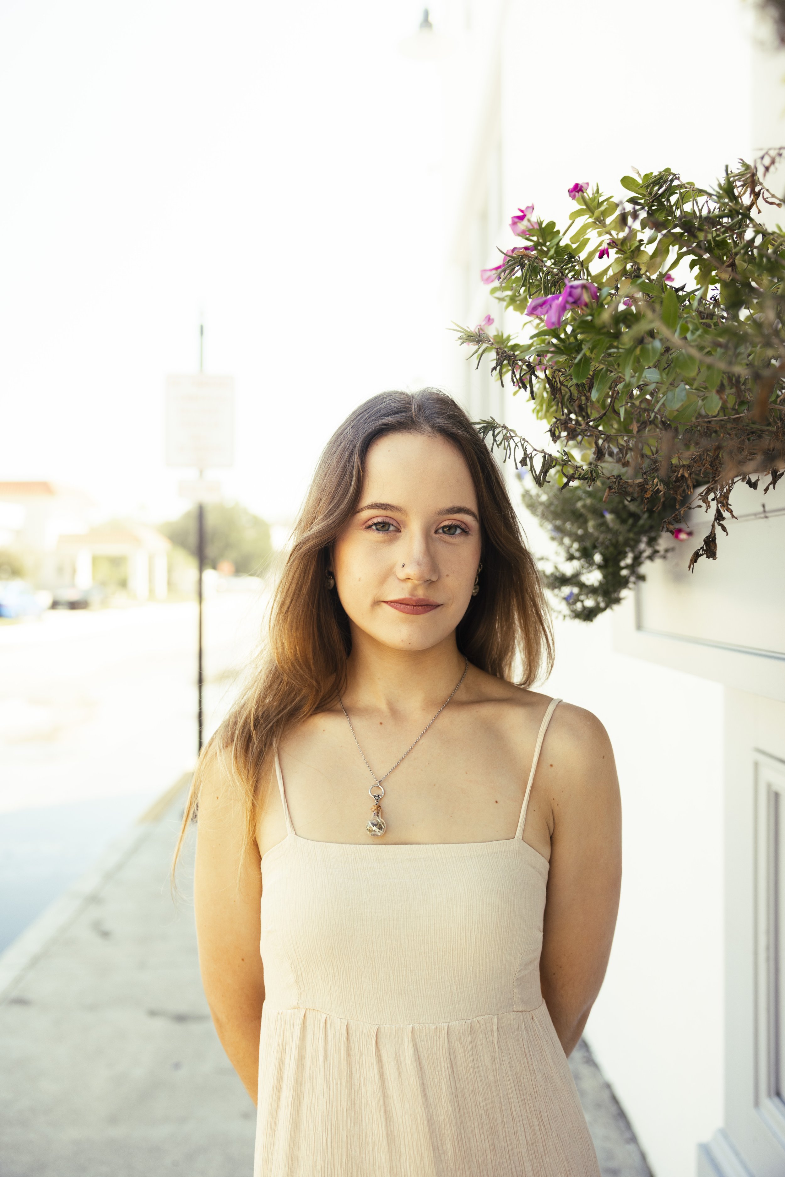 Young woman with light brown hair wearing a beige spaghetti strap dress and necklace standing outdoors near a flowering bush.