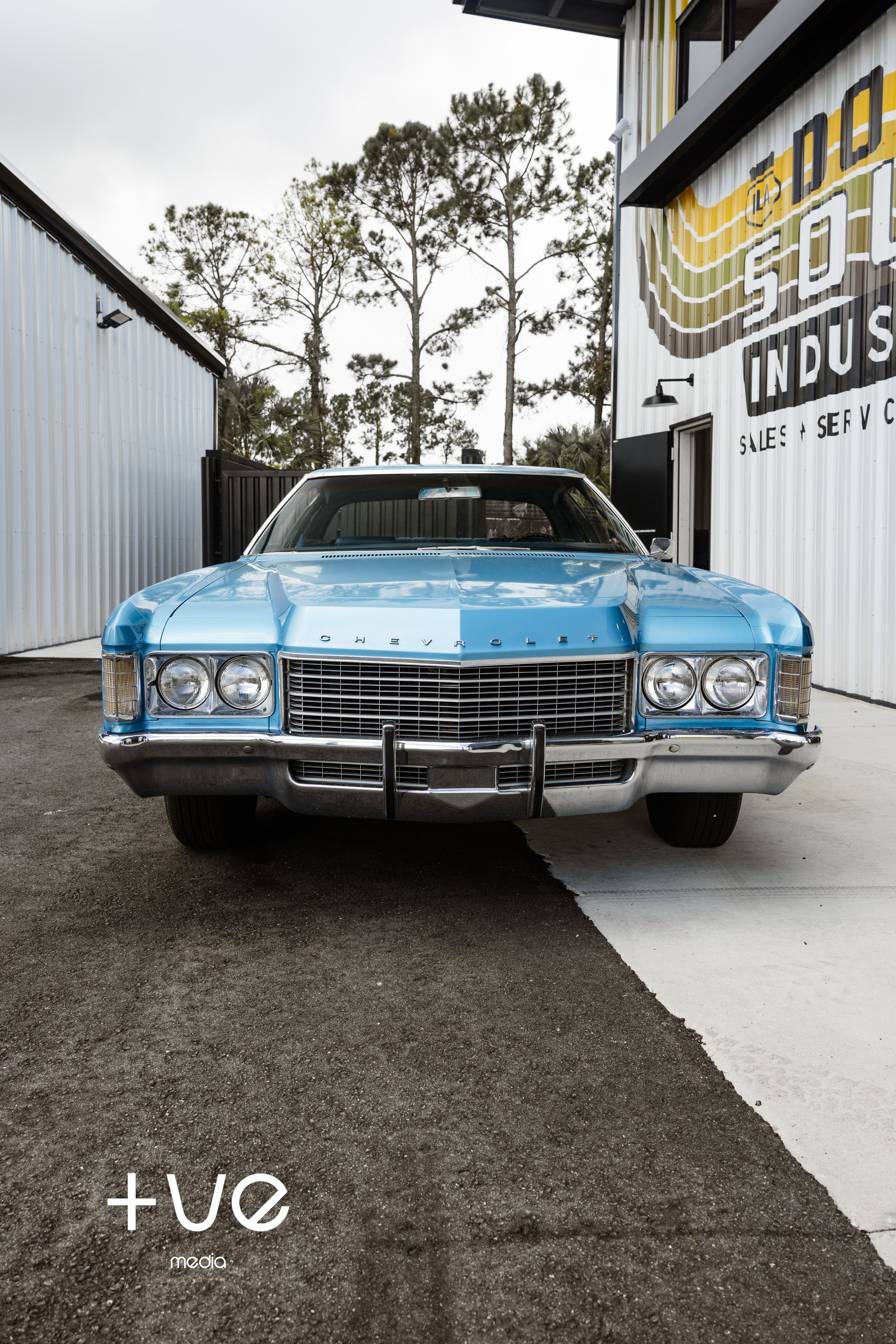 A vintage blue Chevrolet car parked outside a building with sign 'INDUS' and a white metal wall, with trees in the background.