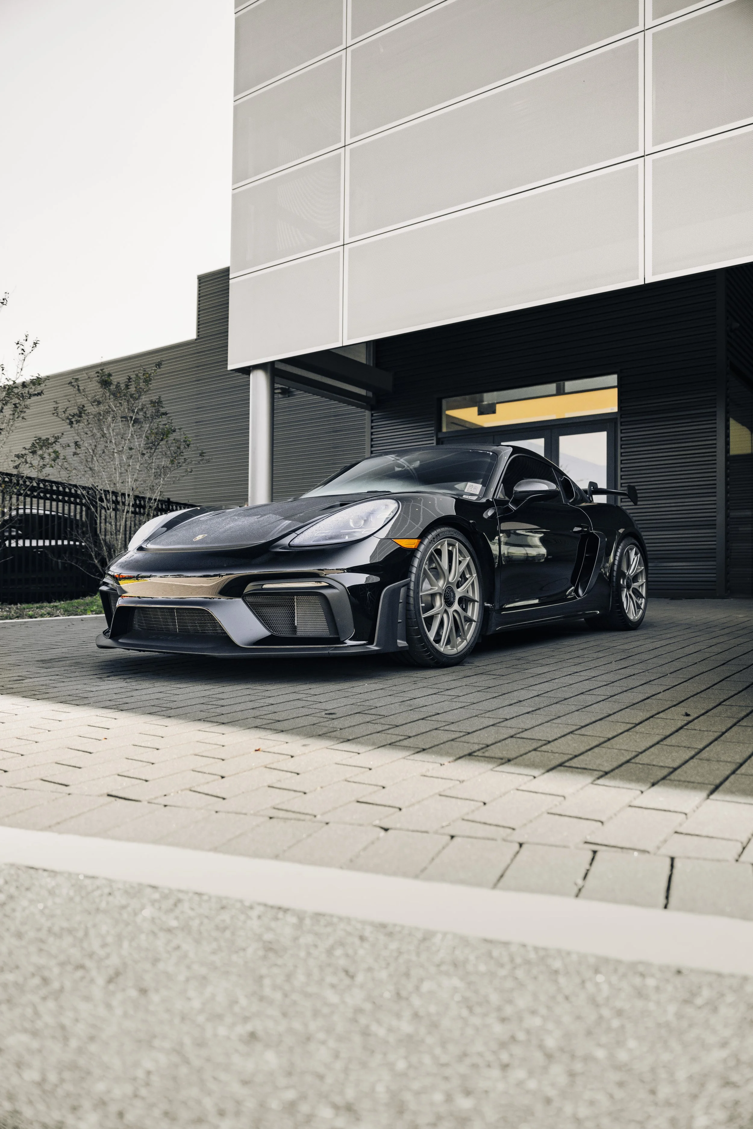 A sleek black Porsche sports car parked outside a modern building with dark exterior walls and large windows.