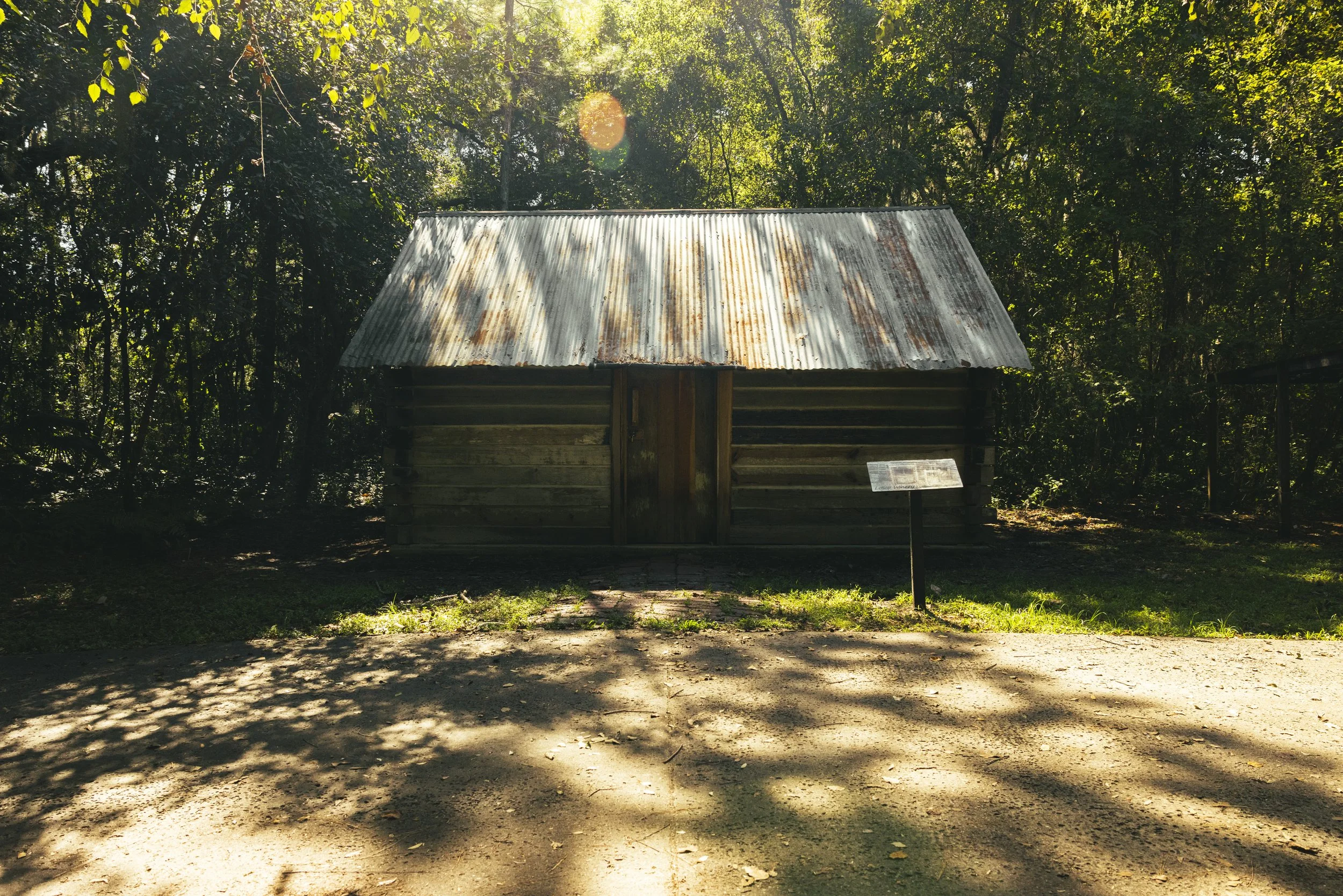 A small rustic wooden cabin with a rusted metal roof, located in a forested area with trees and sunlight casting shadows on the ground.