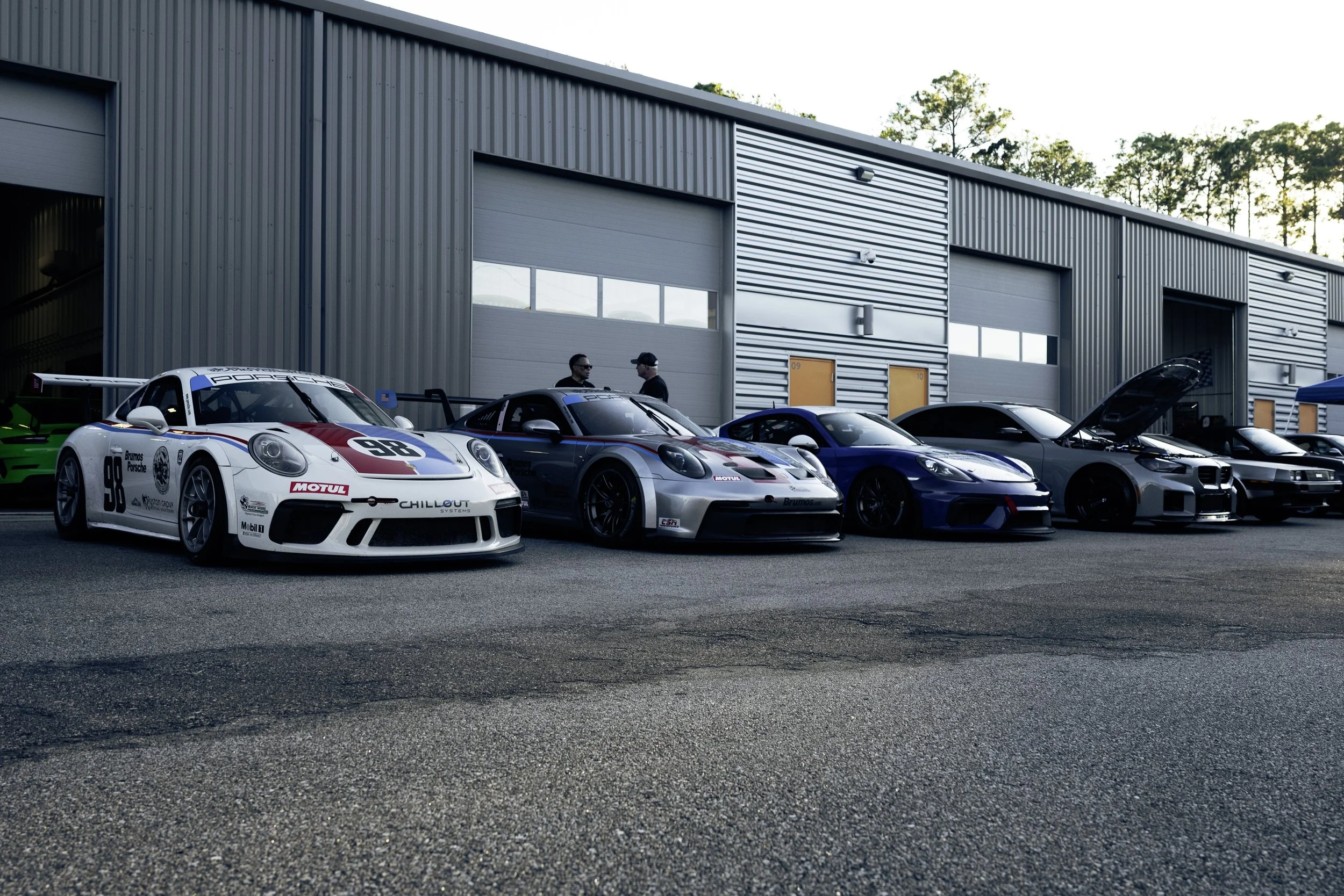 A row of high-performance sports cars parked outside a garage, with two people standing and talking near the cars, and the garage doors open revealing more vehicles inside.