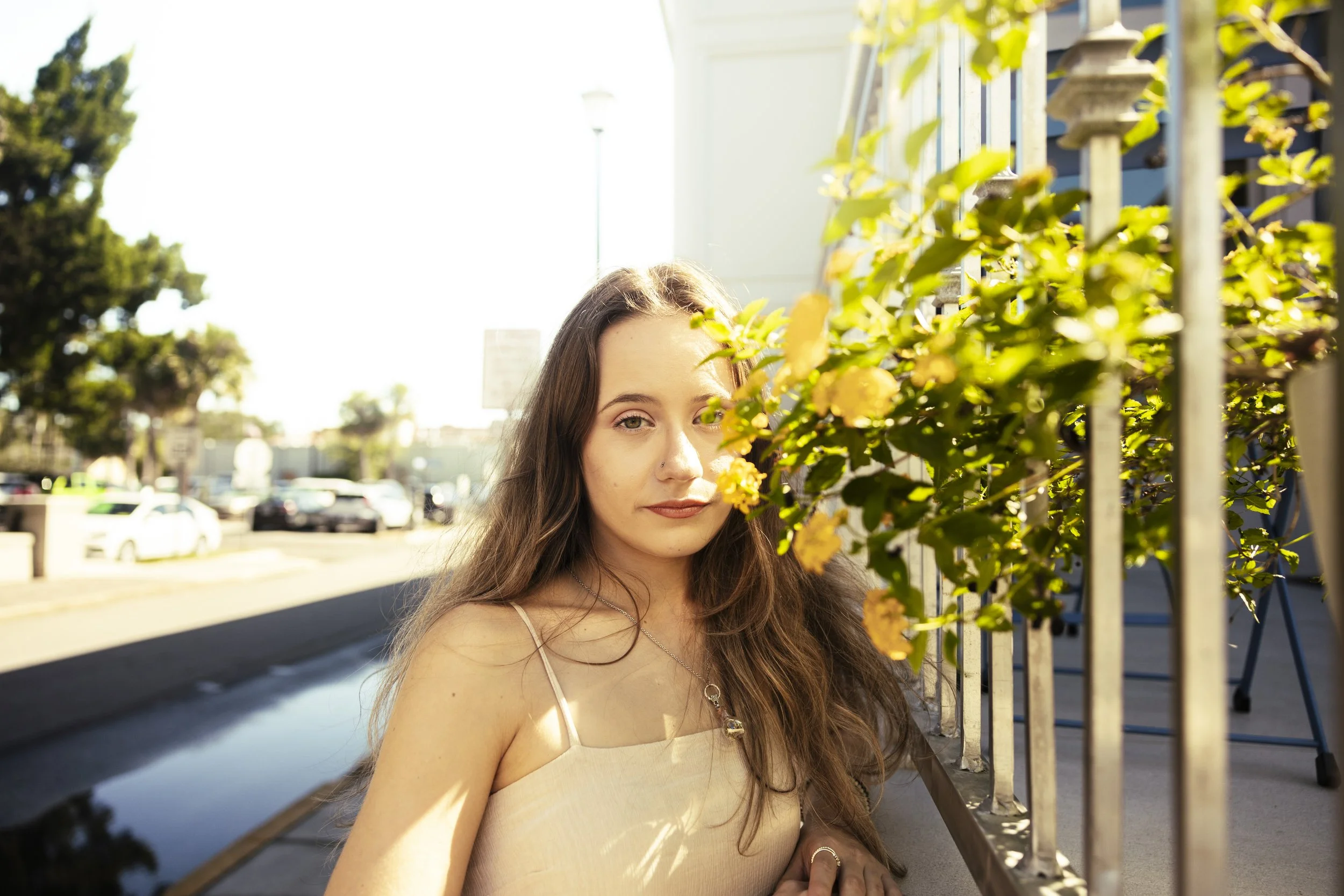 A young woman with long brown hair and fair skin leaning against a wall with yellow flowers and green leaves in front of her, on a sunny street with parked cars and trees in the background.