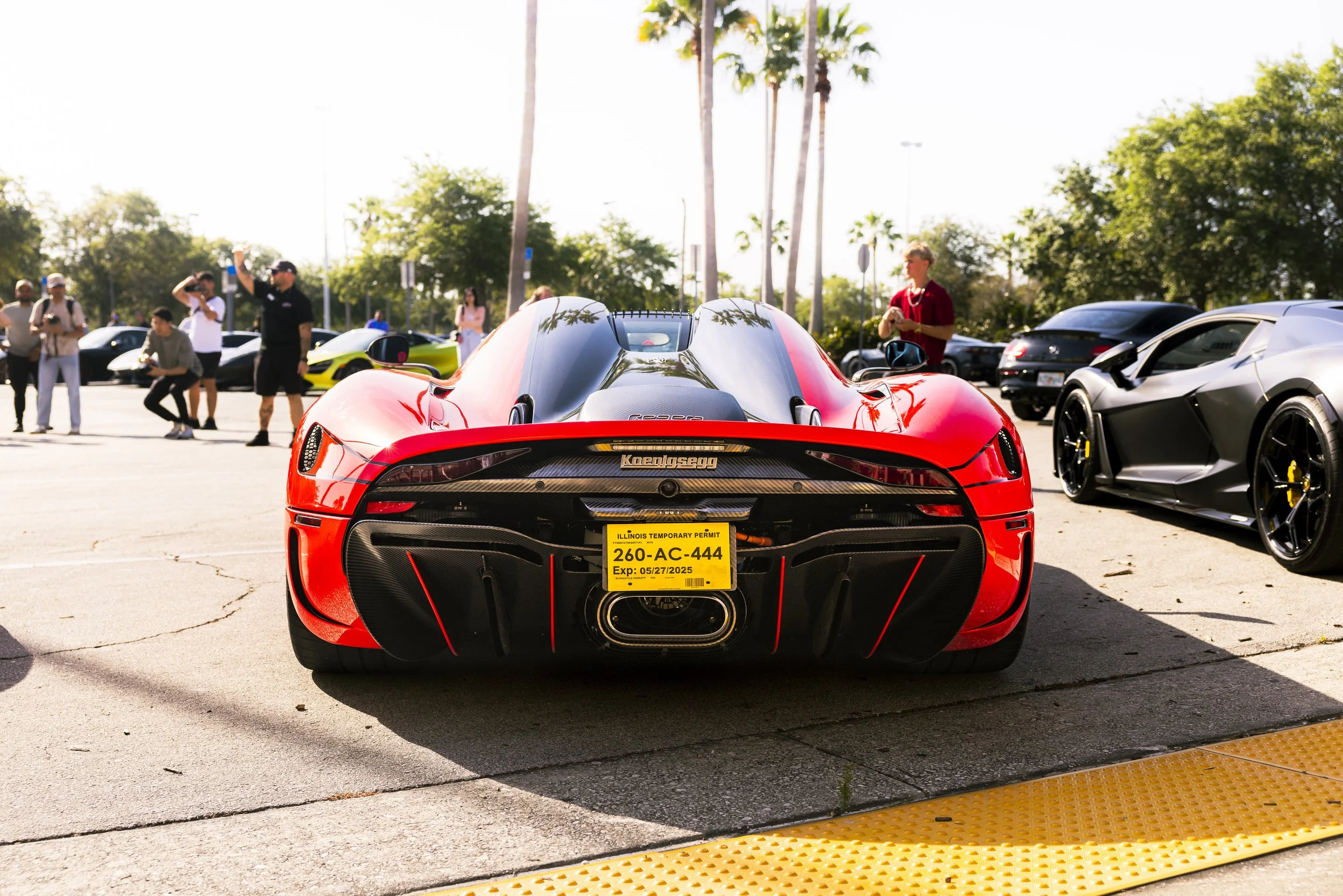 Red Koenigsegg sports car parked among other luxury cars at an outdoor car show, with people admiring and taking photos, palm trees, and blue sky in the background.