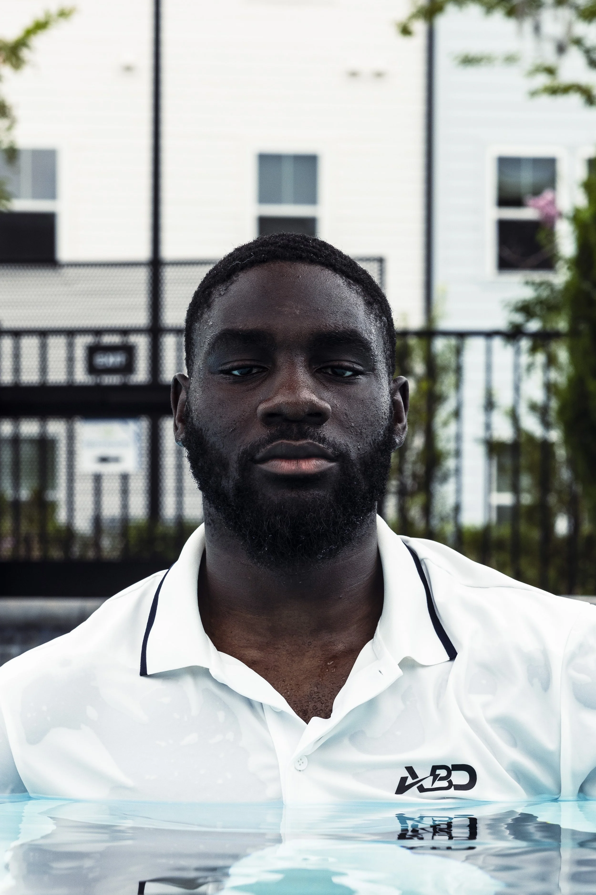 A man with a beard and dark skin wearing a white collared shirt, standing in a pool with water up to his chest, outdoors with a building and trees in the background.