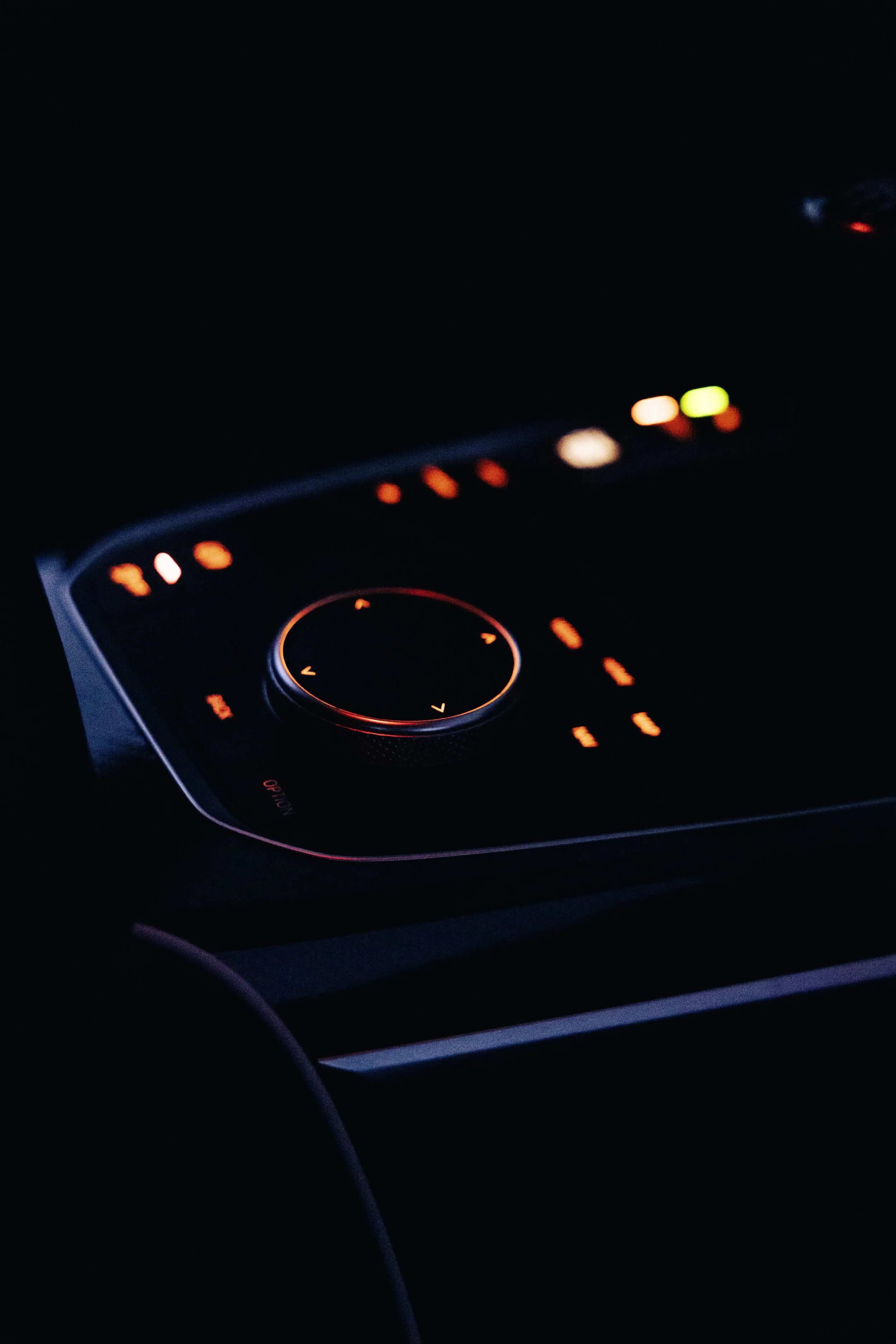 Close-up of a car's illuminated control panel at night with red lights and a dial surrounded by various buttons.