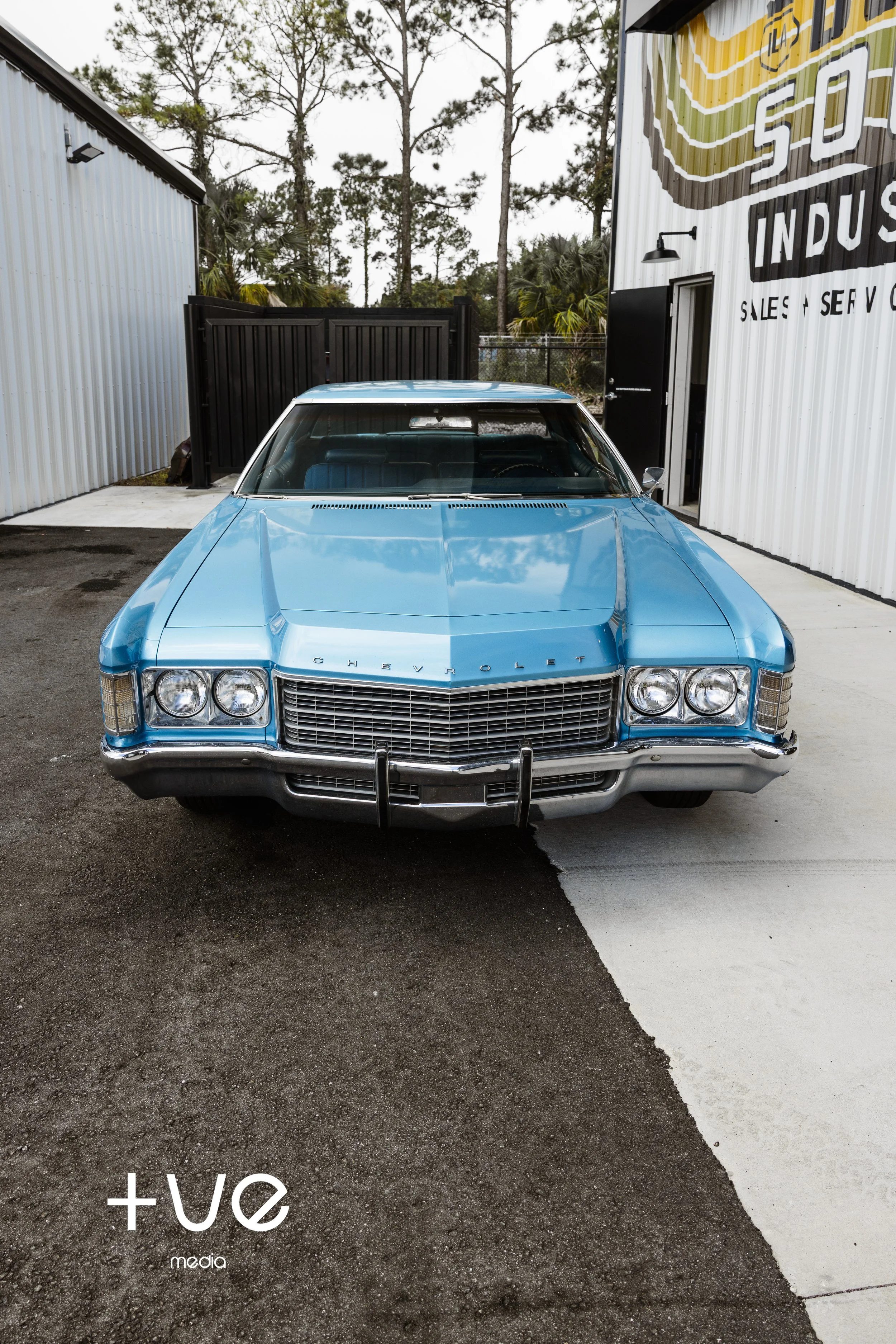 Front view of a classic blue Chevrolet car parked outside a building with white corrugated metal siding, black fence, and trees in the background.