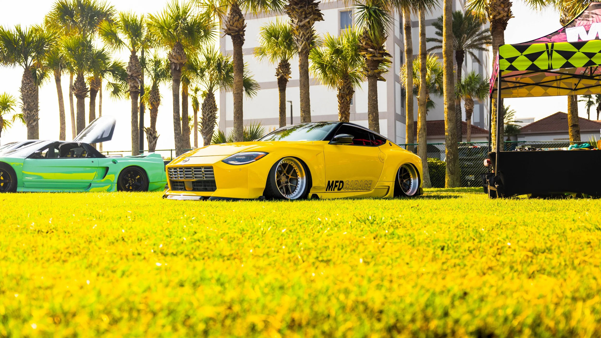 A yellow sports car and a green and black race car parked on a grassy field with palm trees and a white building in the background during daytime.