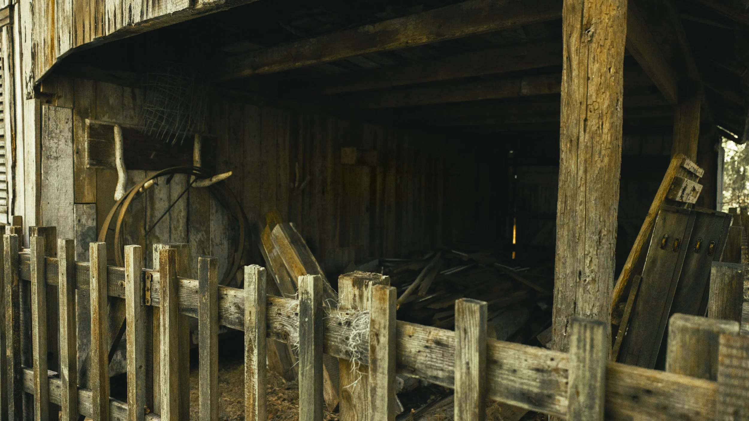 Interior of an old, abandoned wooden shed with damaged walls and debris on the floor, viewed through a broken wooden fence.