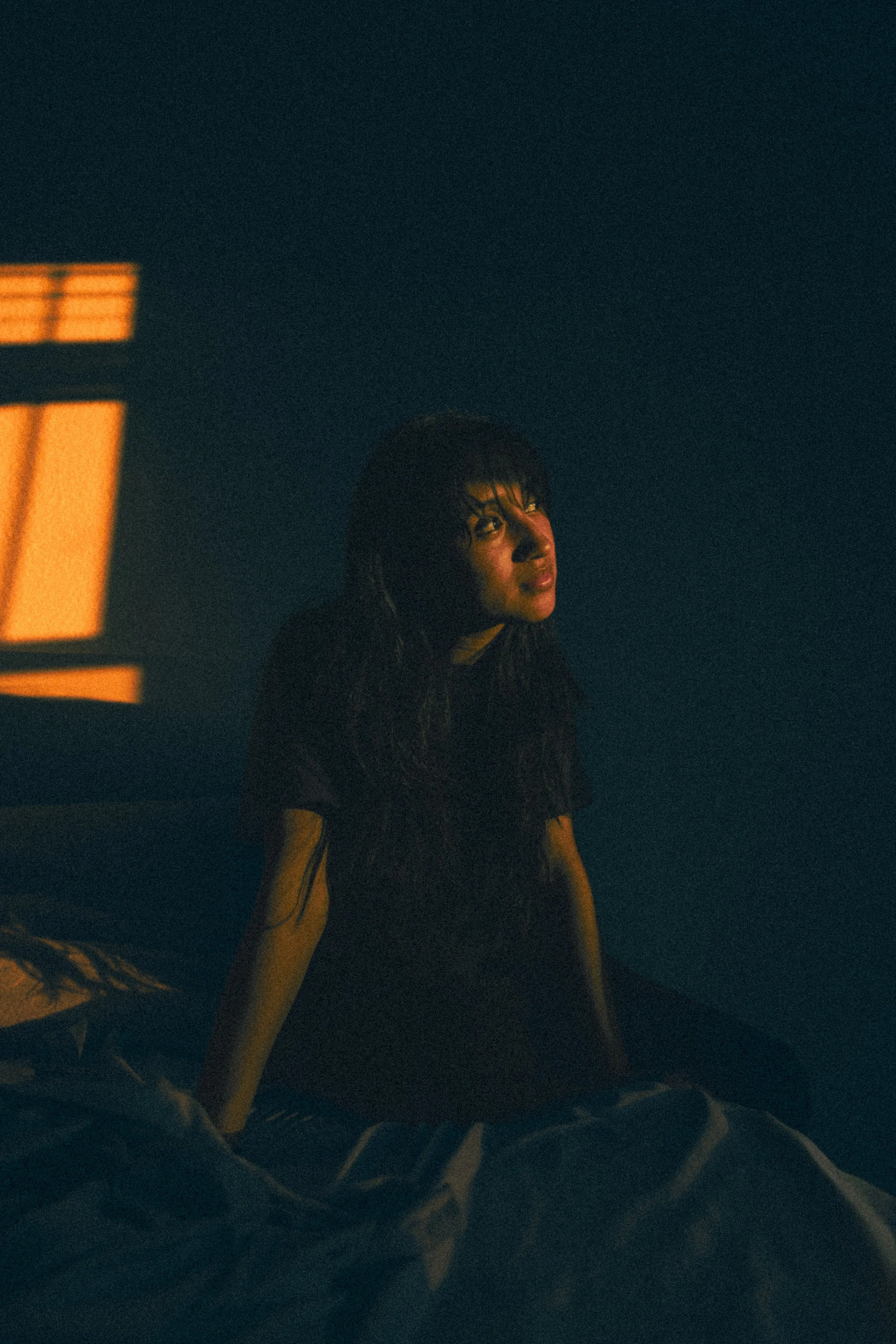 A woman with wet hair sitting on a bed in a dark room illuminated by a warm orange light coming through a window.