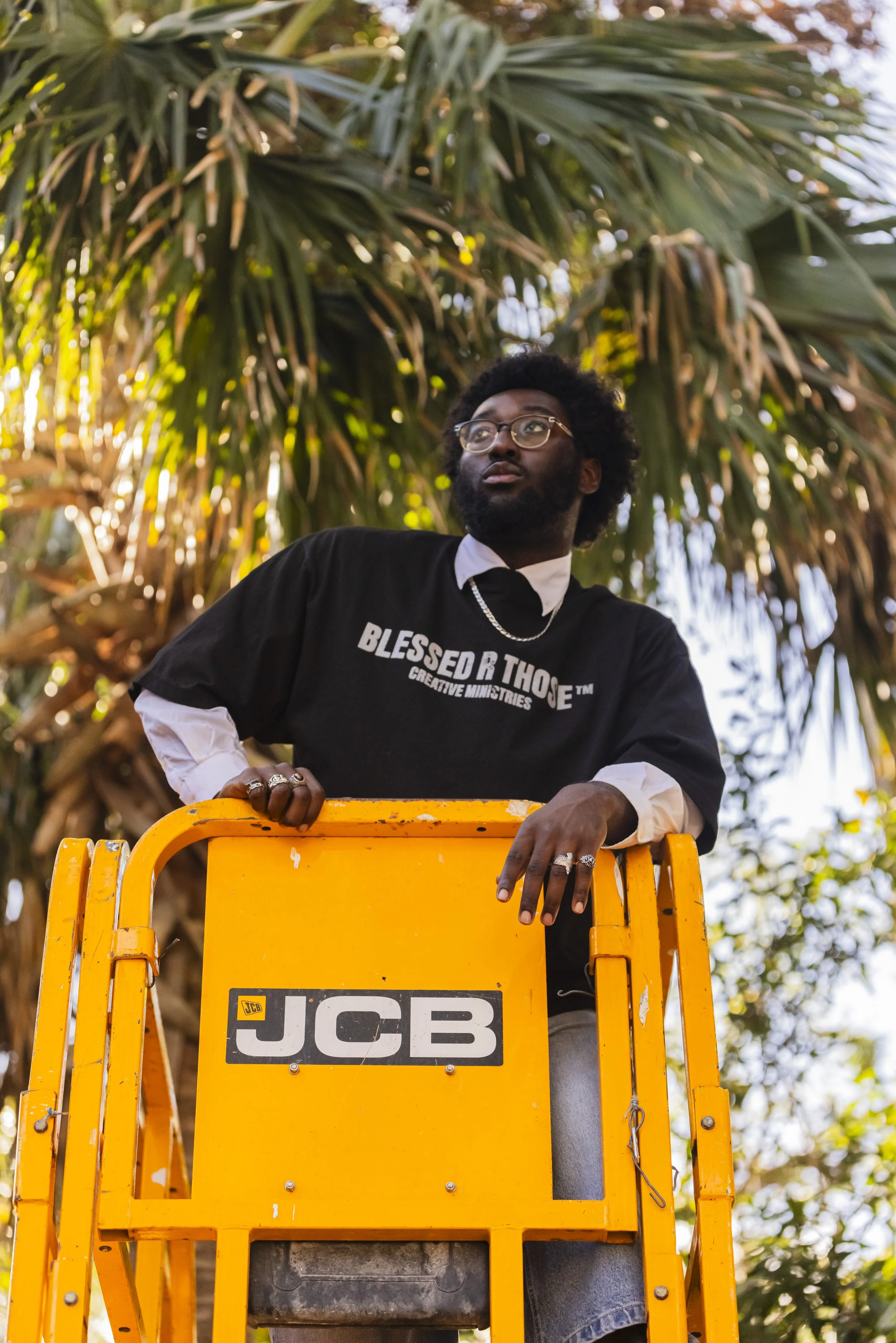 A man with glasses and a beard standing on a yellow JCB lift, looking thoughtfully into the distance, with a large leafy tree in the background.