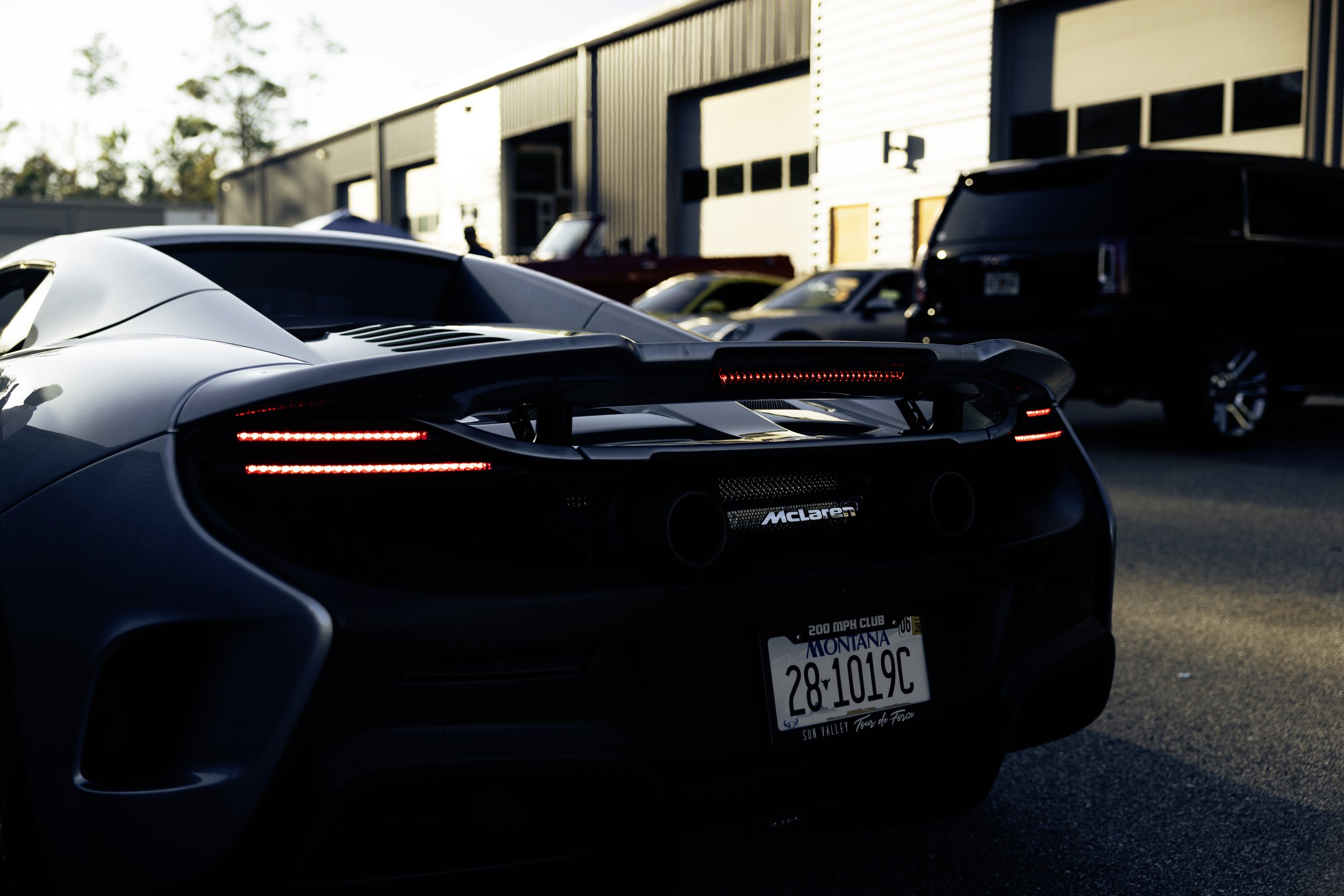 The rear of a silver McLaren sports car with custom Montana license plate, parked outside a building with other vehicles including a black SUV and other cars.