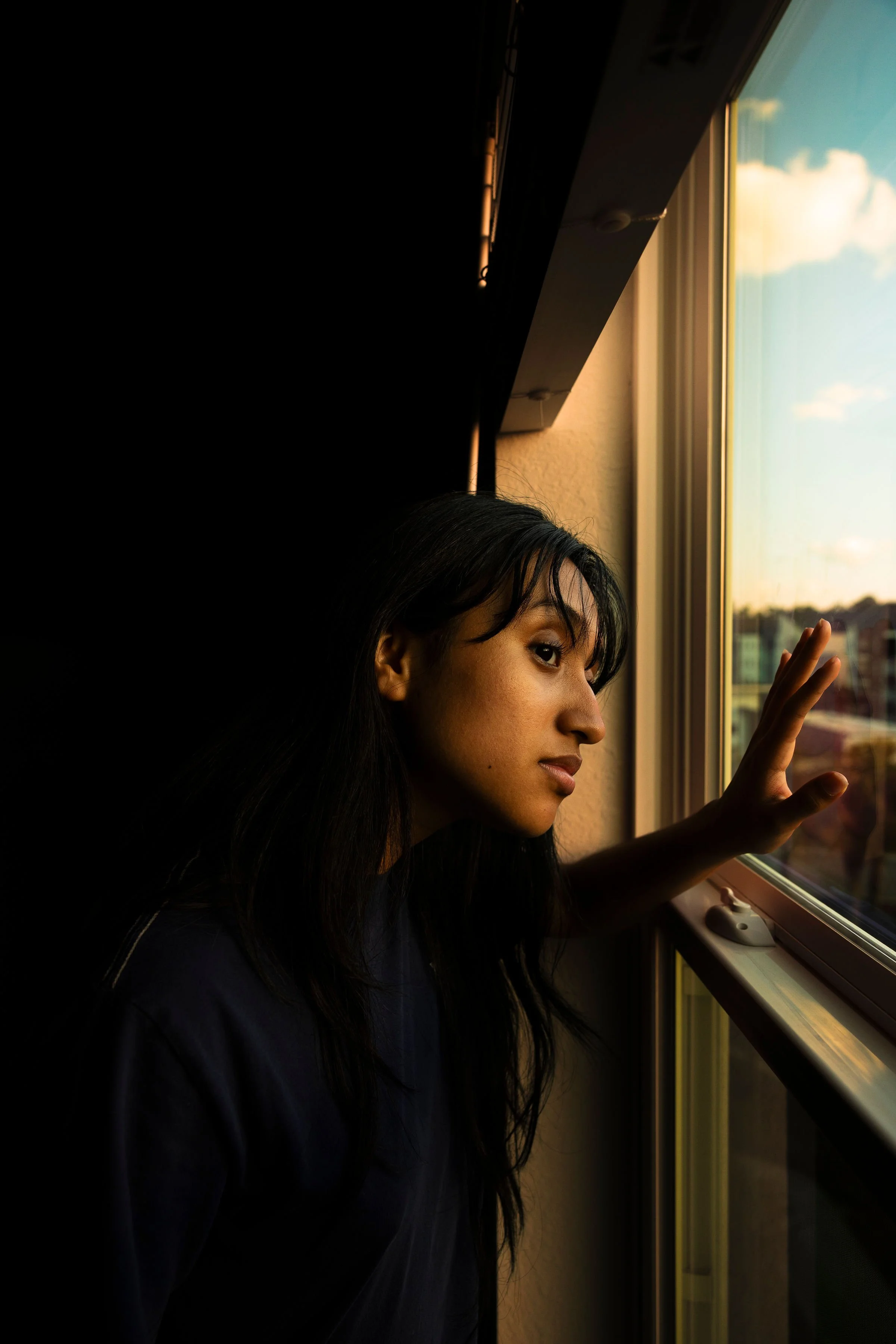 A woman with dark hair looking out a window during sunset, with her hand on the glass.