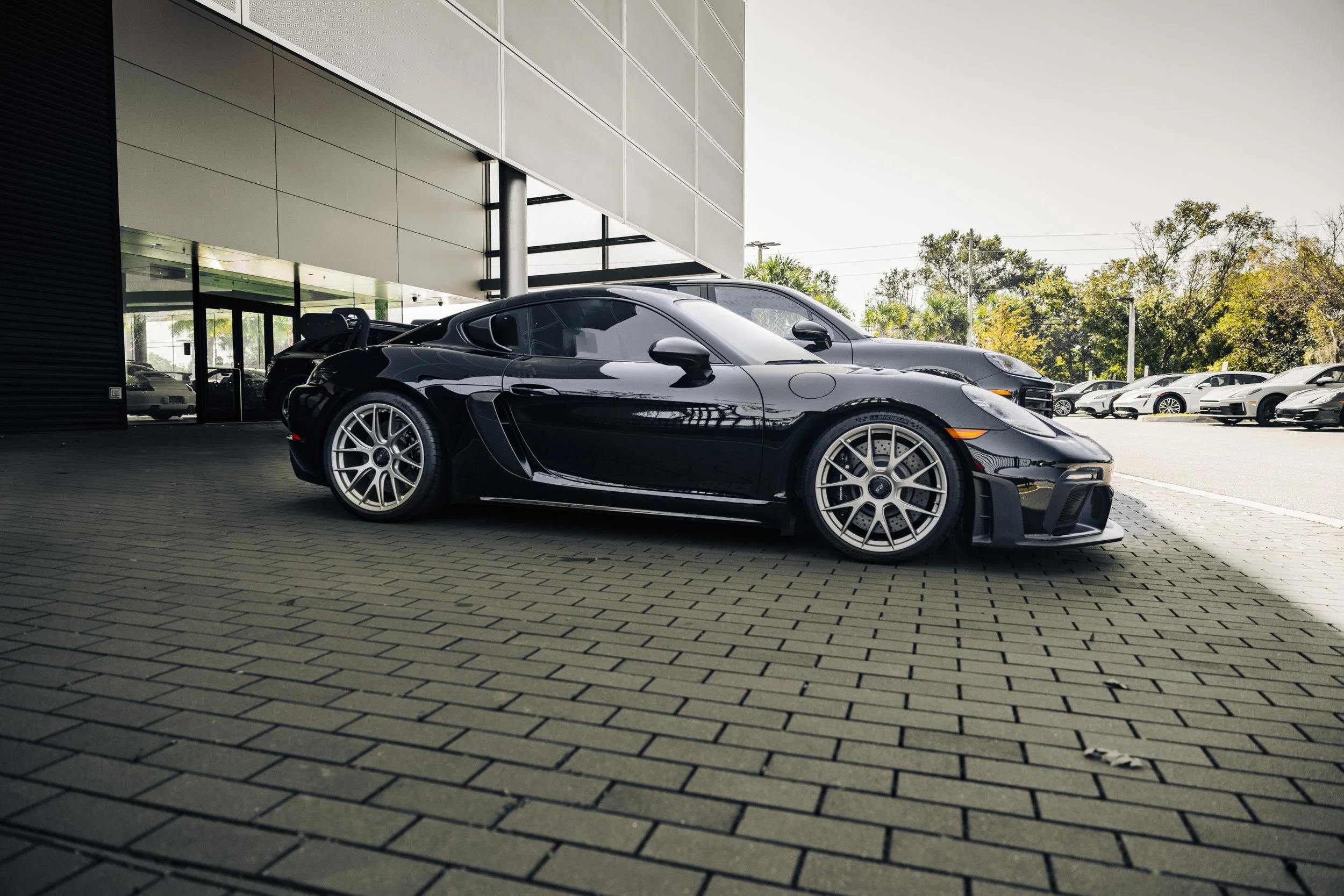 Black sports car, likely a Porsche 718 Cayman, parked outside a modern building with large glass windows, other cars visible in the background, and a cloudy sky.