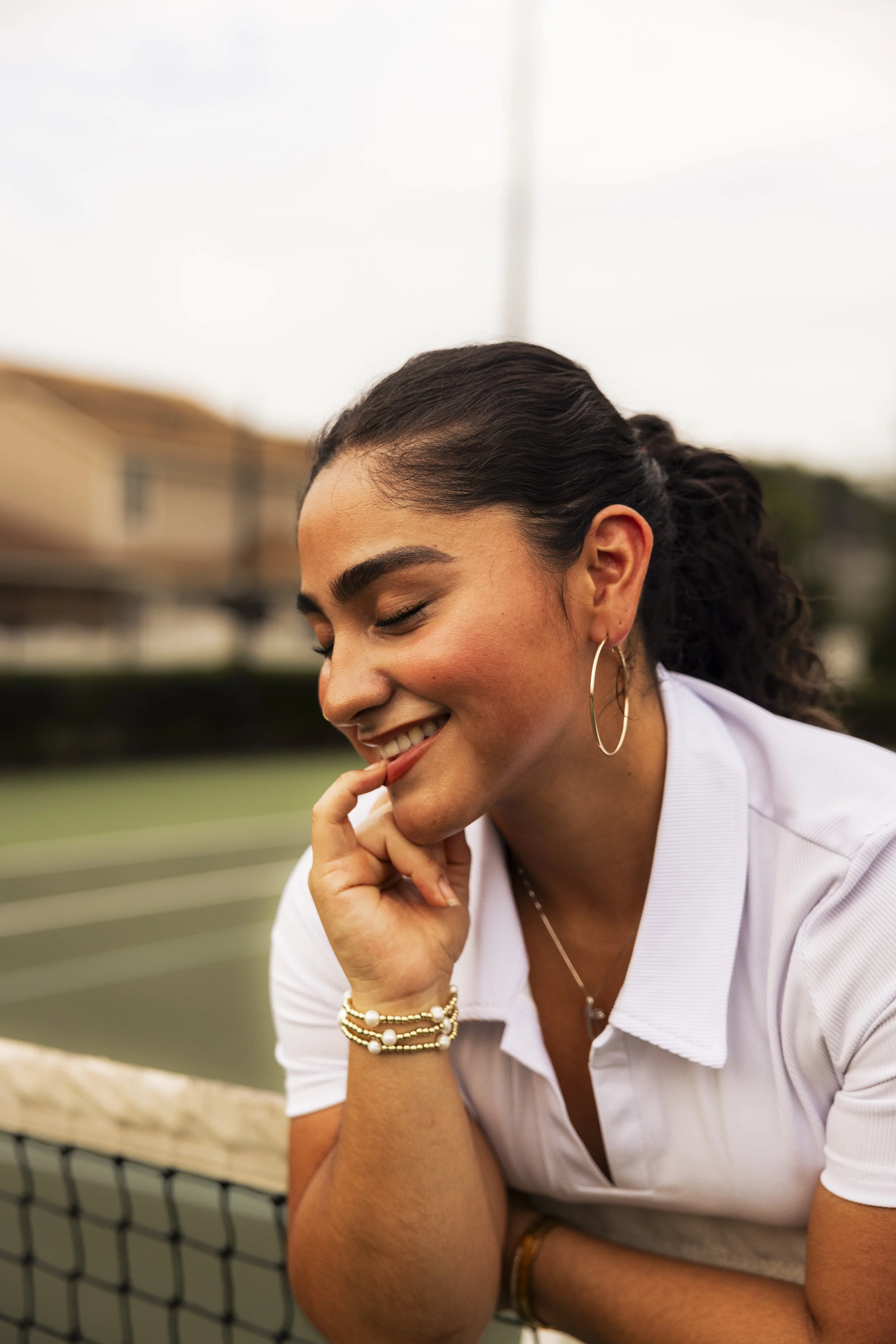 A young woman with dark curly hair, wearing hoop earrings, a white polo shirt, and jewelry, smiles with her eyes closed while resting her chin on her hand on a tennis court.