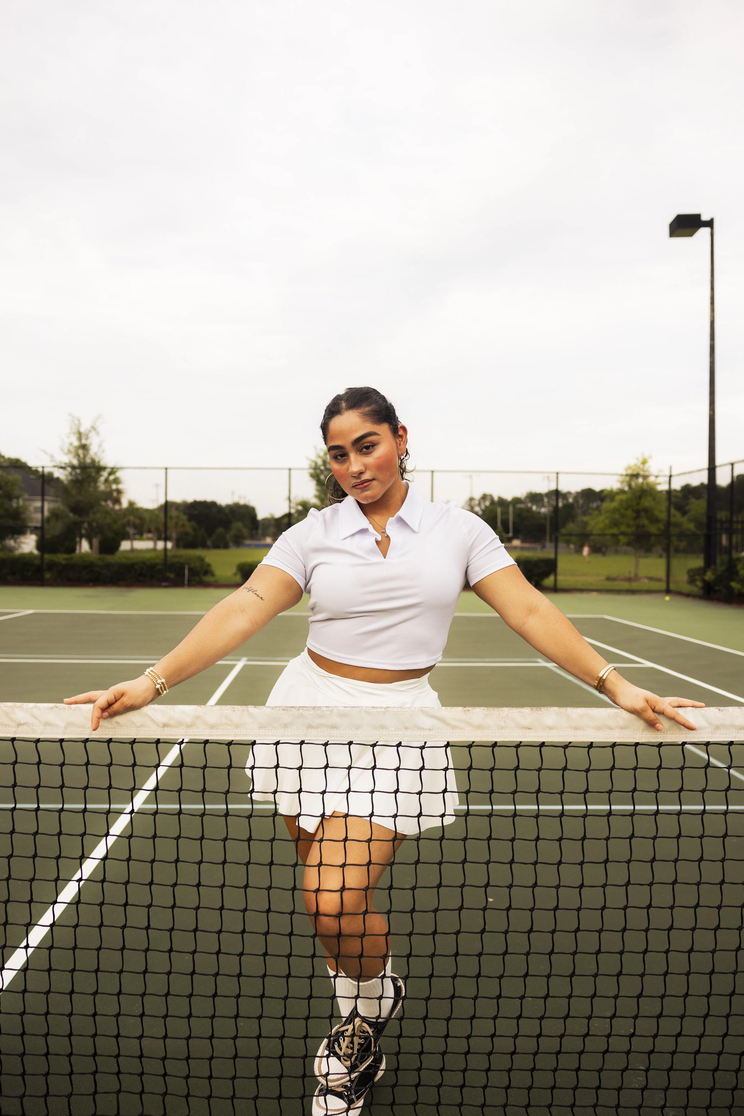 A woman standing on a tennis court, leaning on the net, wearing a white crop top and tennis skirt, with a cloudy sky in the background.