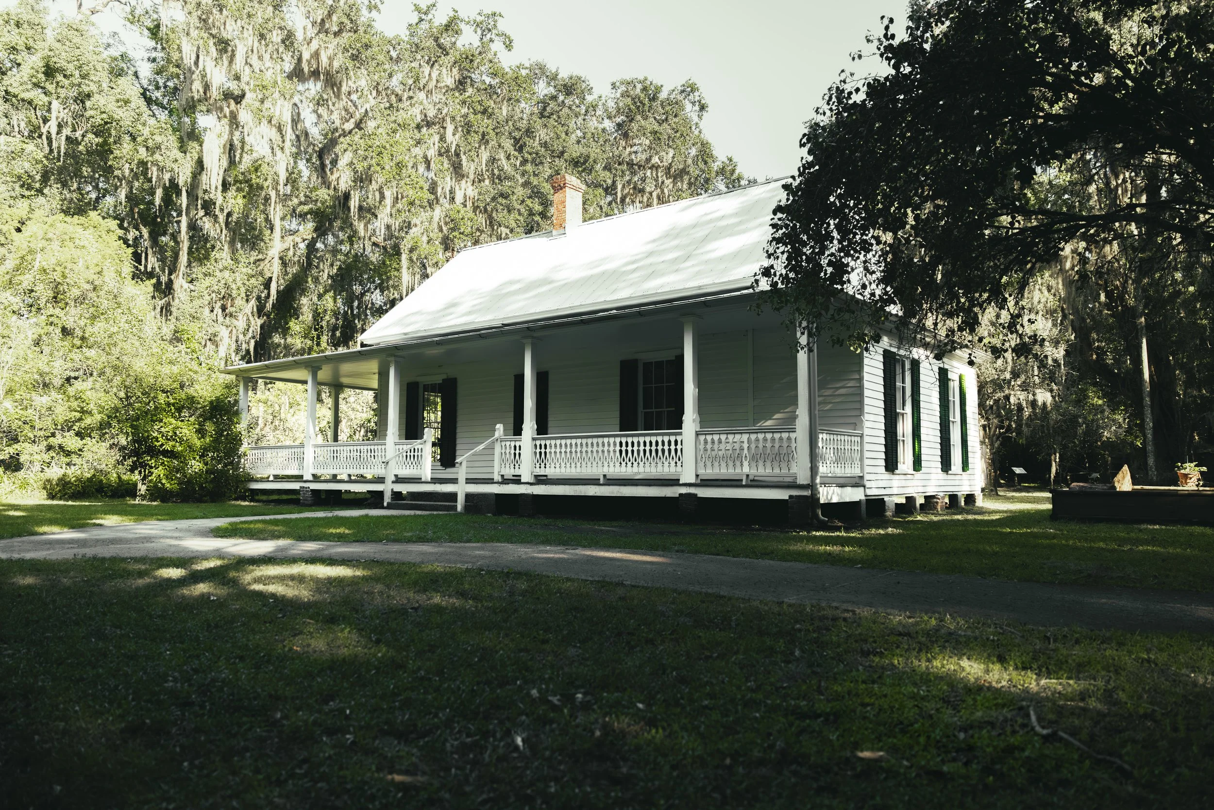 A historic white house with black shutters and a metal roof, surrounded by lush greenery and trees, with a porch and a driveway in the front.