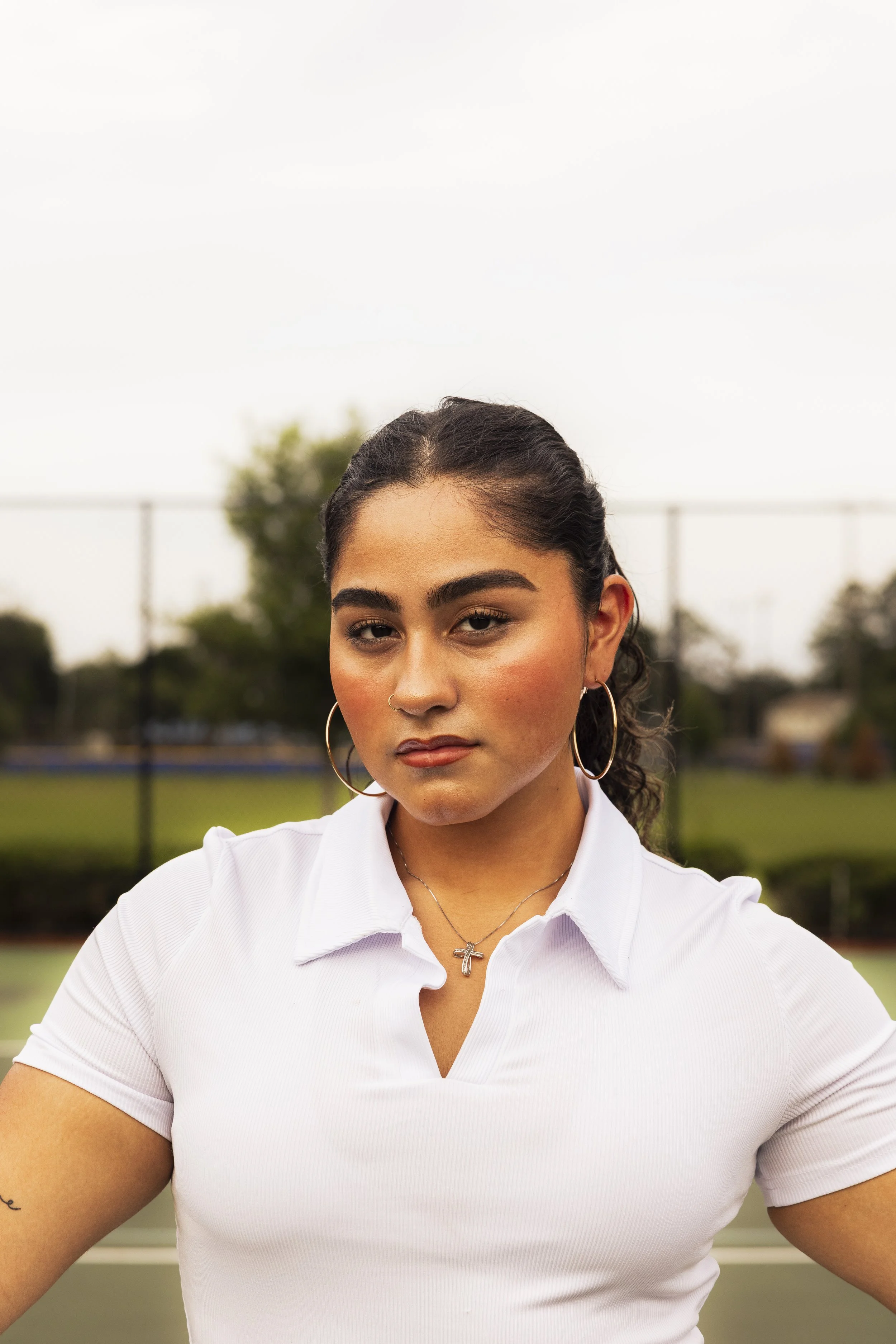 A young woman with dark, curly hair wearing hoop earrings, a cross necklace, and a white polo shirt stands outdoors on a tennis court with trees and a fence in the background.