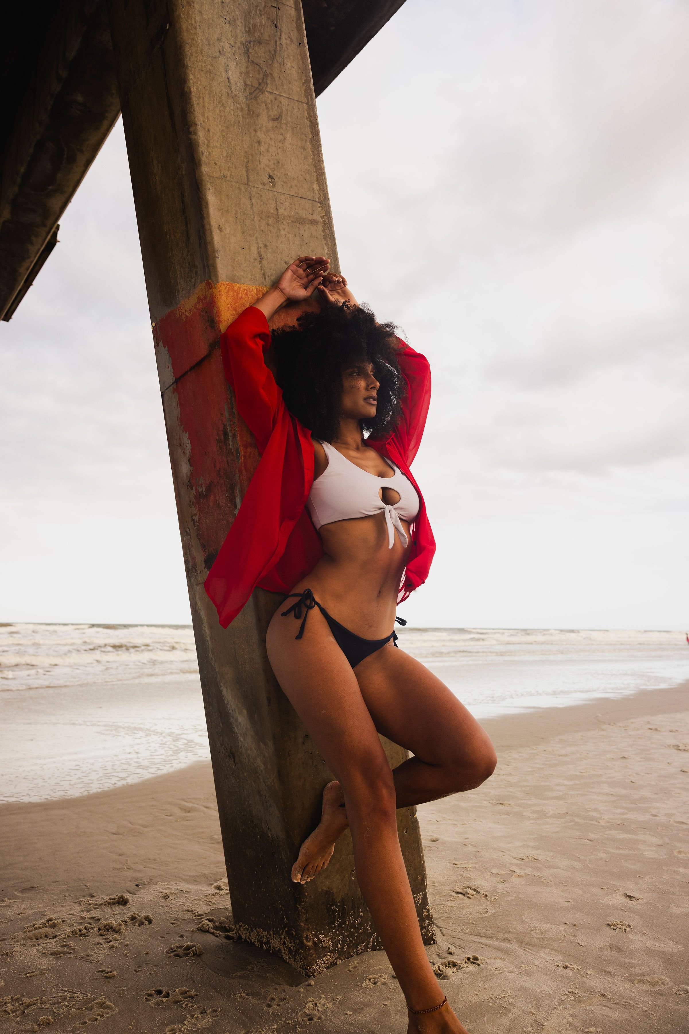 A woman with curly hair leaning against a large concrete pillar on a beach, wearing a white bikini top with a knot and cut-out design, black bikini bottoms, and a red cover-up. The beach and ocean are visible in the background under an overcast sky.