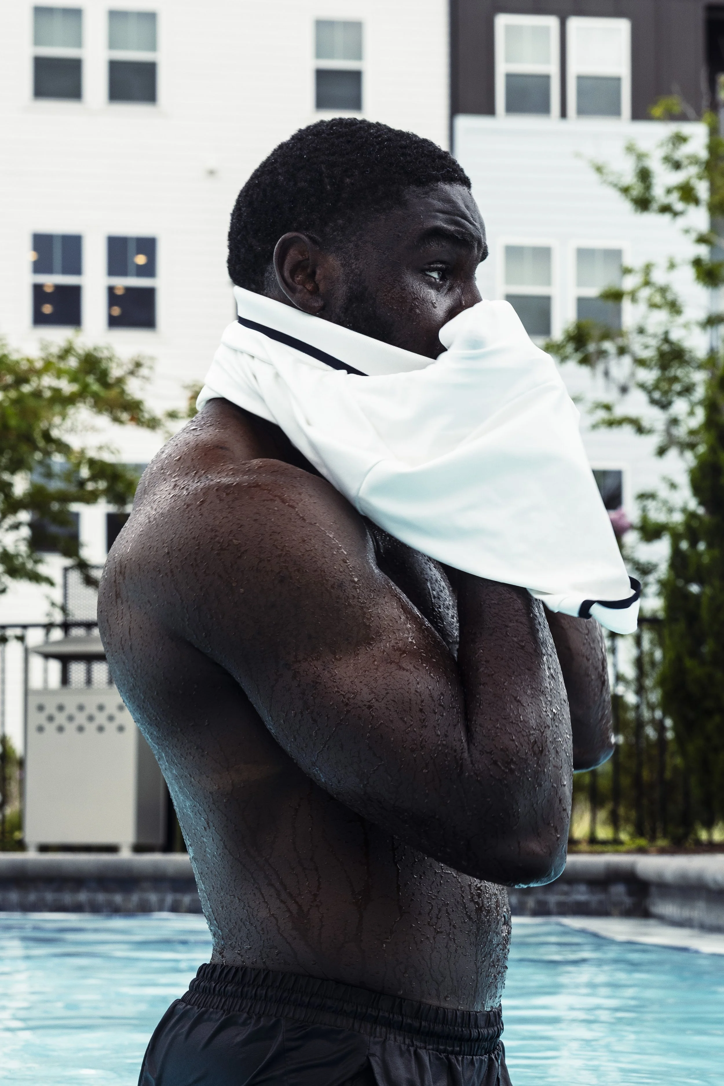 A man with wet skin standing at a swimming pool, holding a white shirt around his neck and covering part of his face, with residential buildings in the background.