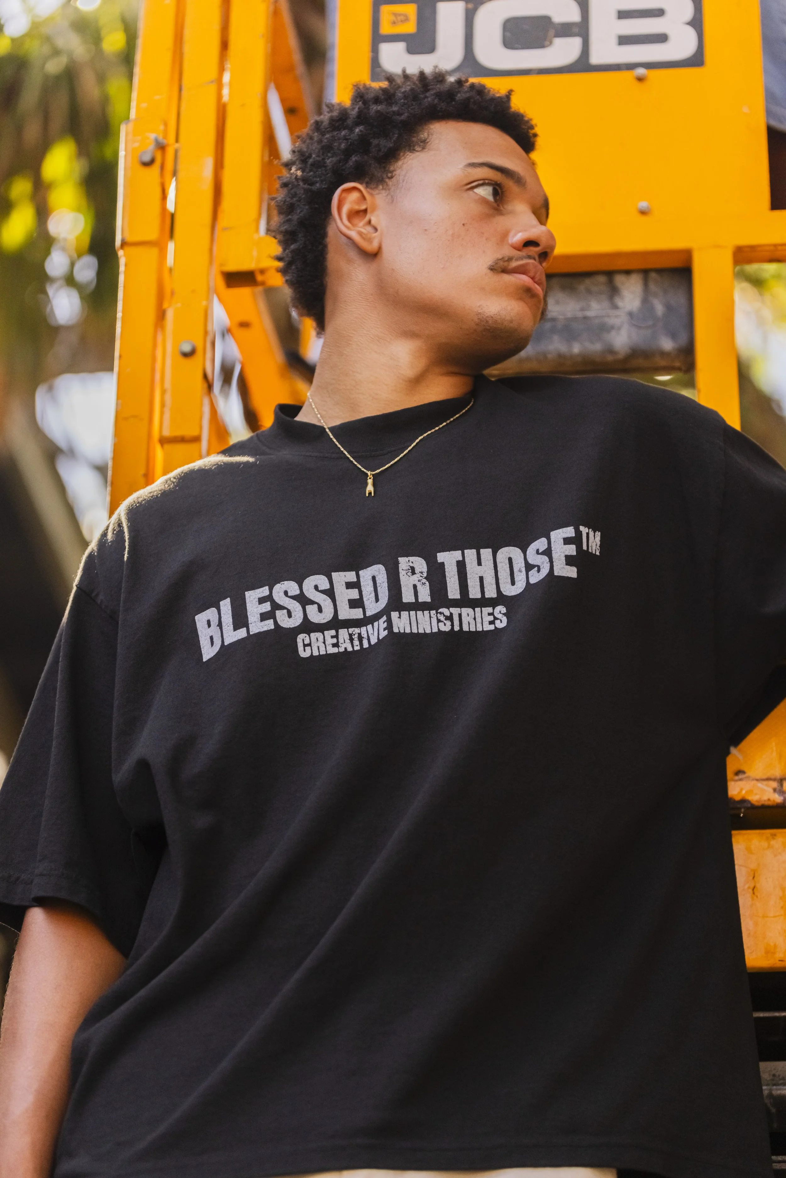 A young man with curly hair wearing a black t-shirt that reads "BLESSED R THOSE™ CREATIVE MINISTRIES" stands in front of an orange construction lift with "JCB" branding.