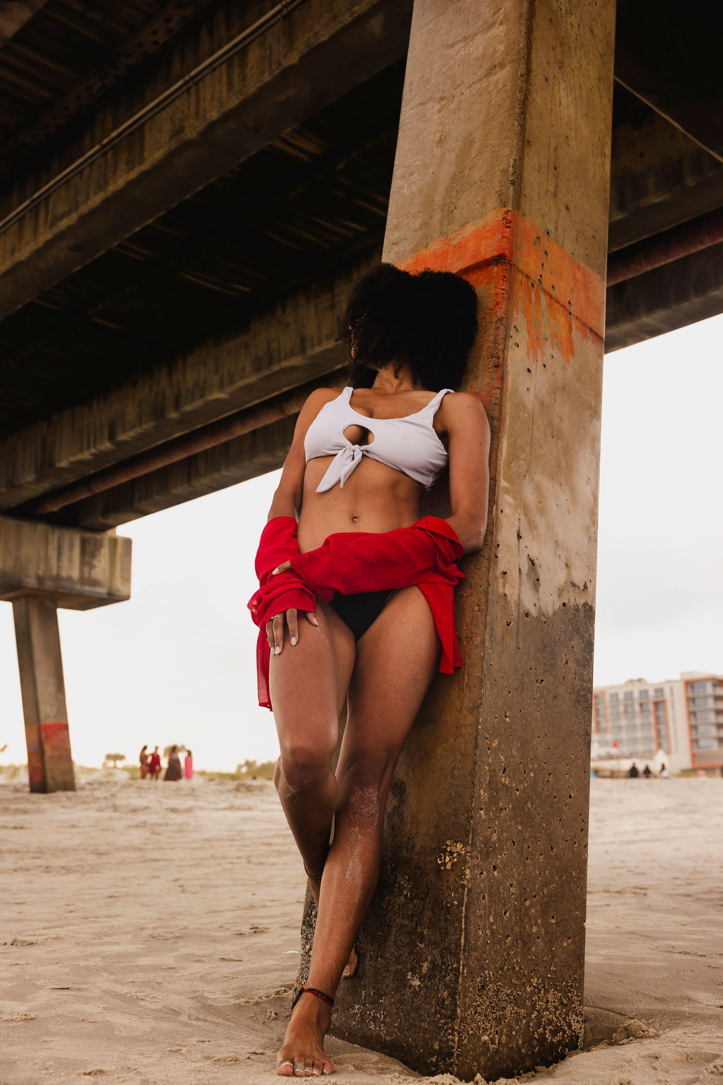 A woman wearing a white crop top, black underwear, and a red shirt draped around her waist, standing barefoot on a sandy beach and leaning against a large concrete pillar under a bridge.