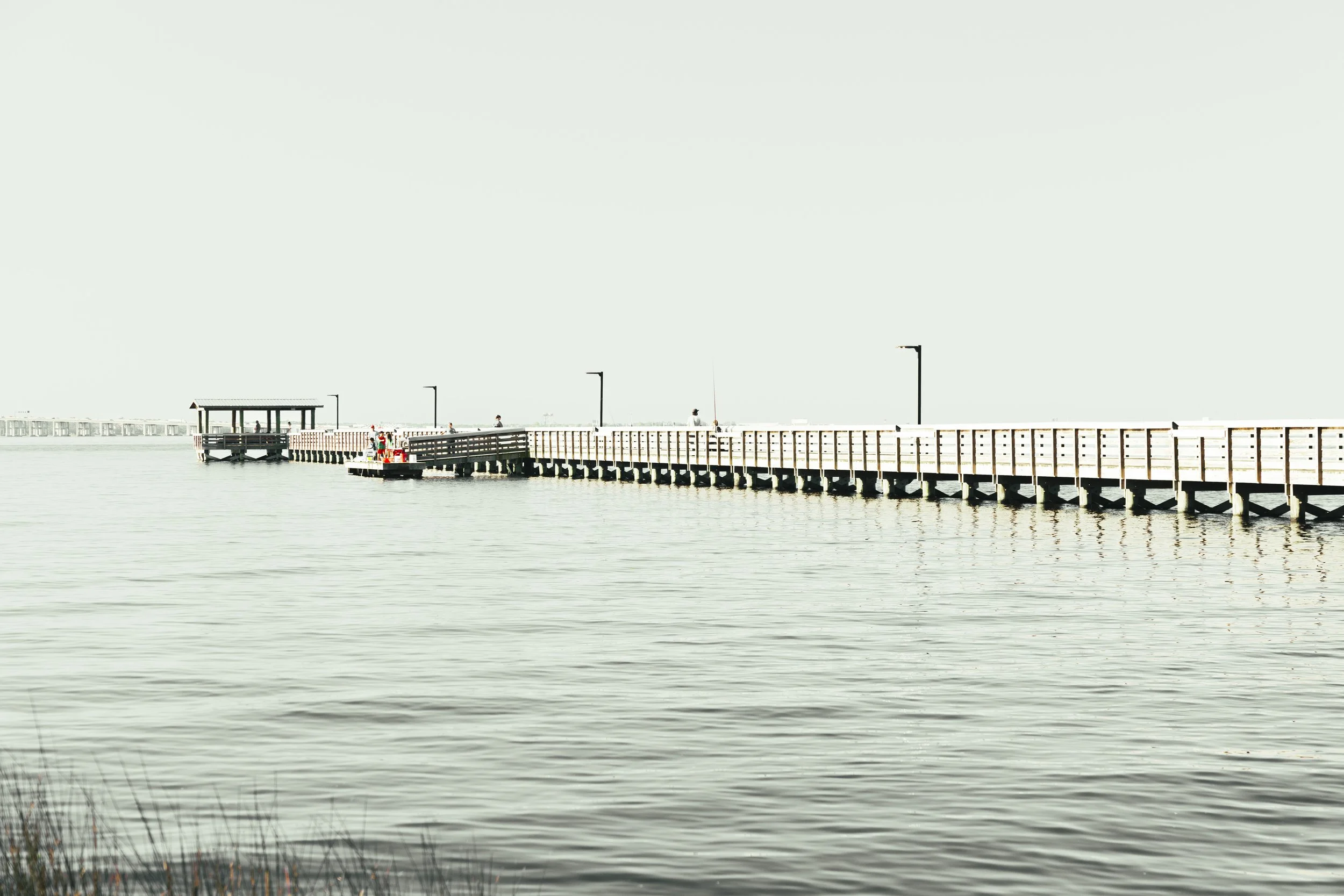 A long wooden pier extends over a calm body of water, with a small shelter at the end and several people walking along it; the sky is bright and clear.