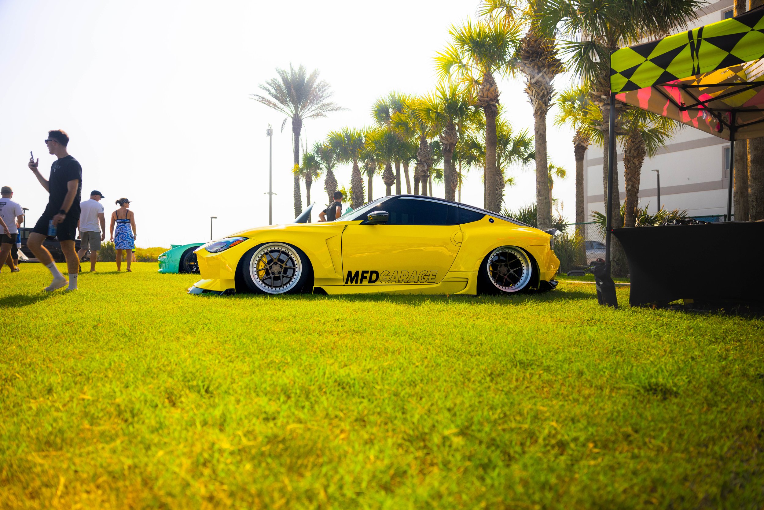 Yellow sports car with lowered stance and custom wheels parked on grass at outdoor car show, with palm trees and people in background.