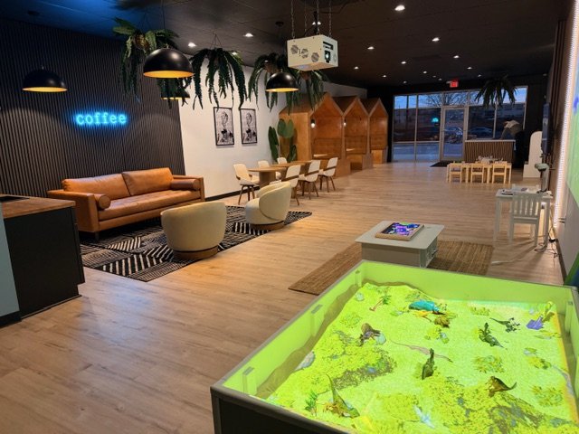 Coffee shop with lounge area, dining tables, children's play area with a water table, and decorated with hanging plants.
