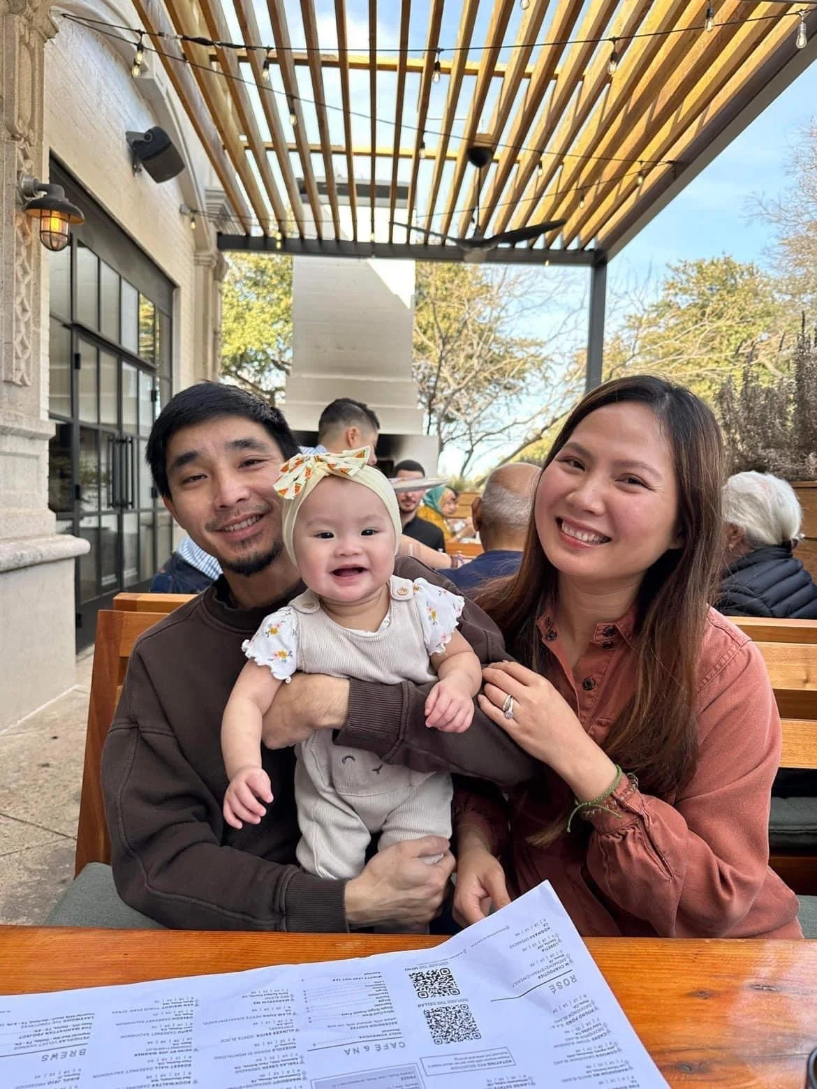 A family of three, a man, a woman, and a baby, sitting at an outdoor restaurant enjoying a moment together with trees and other patrons in the background.