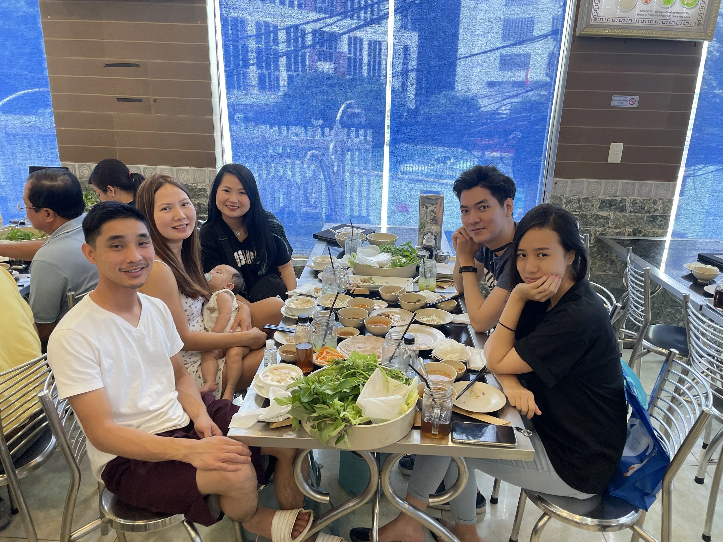 A group of seven people, including a baby, sitting around a table in a restaurant after a meal, with plates of food, bowls, drinks, and vegetables on the table.