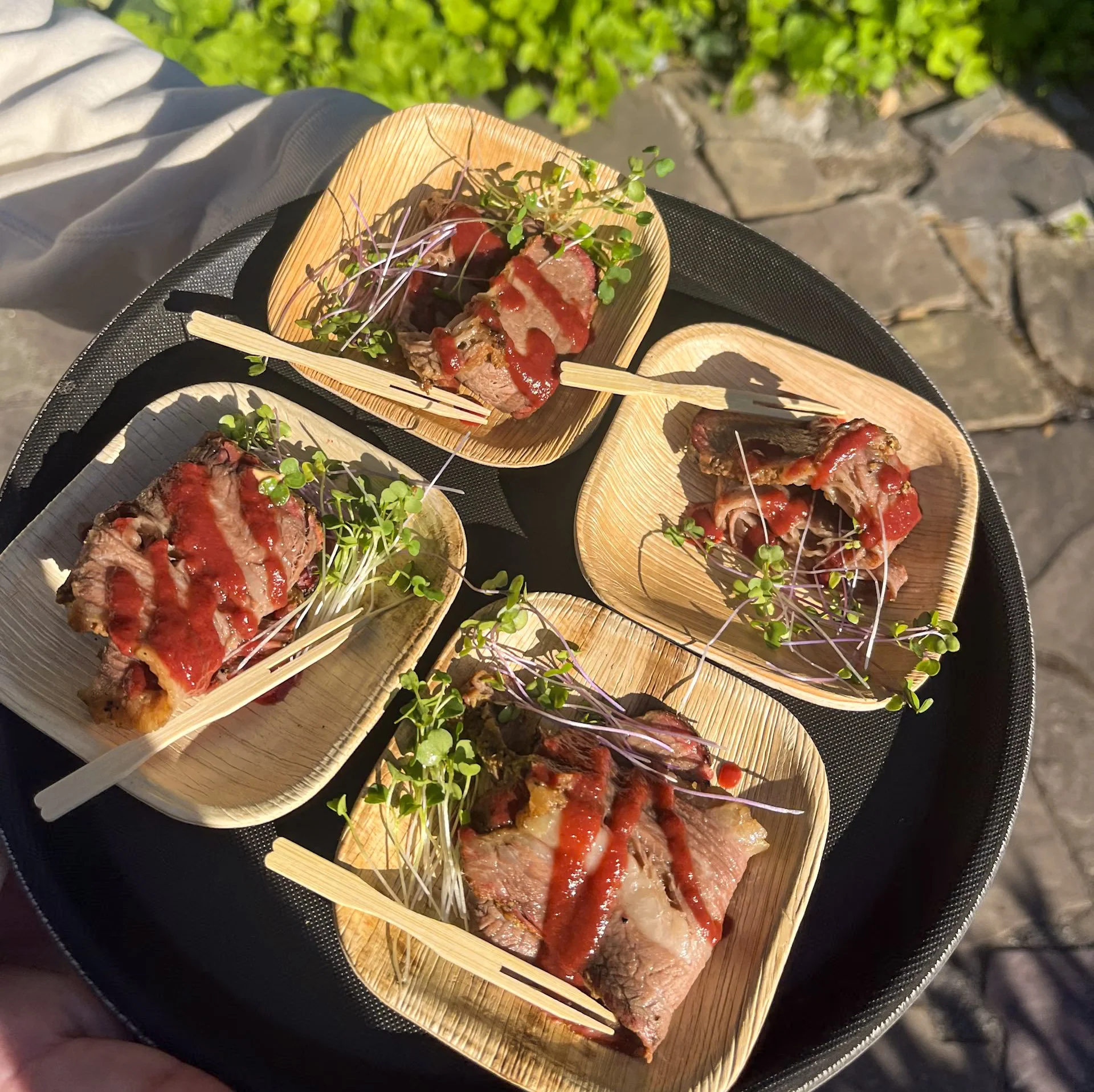slices of smoked brisket with a red sauce and microgreens on wooden plates with small wooden forks on a black tray with tile and greenery in the background