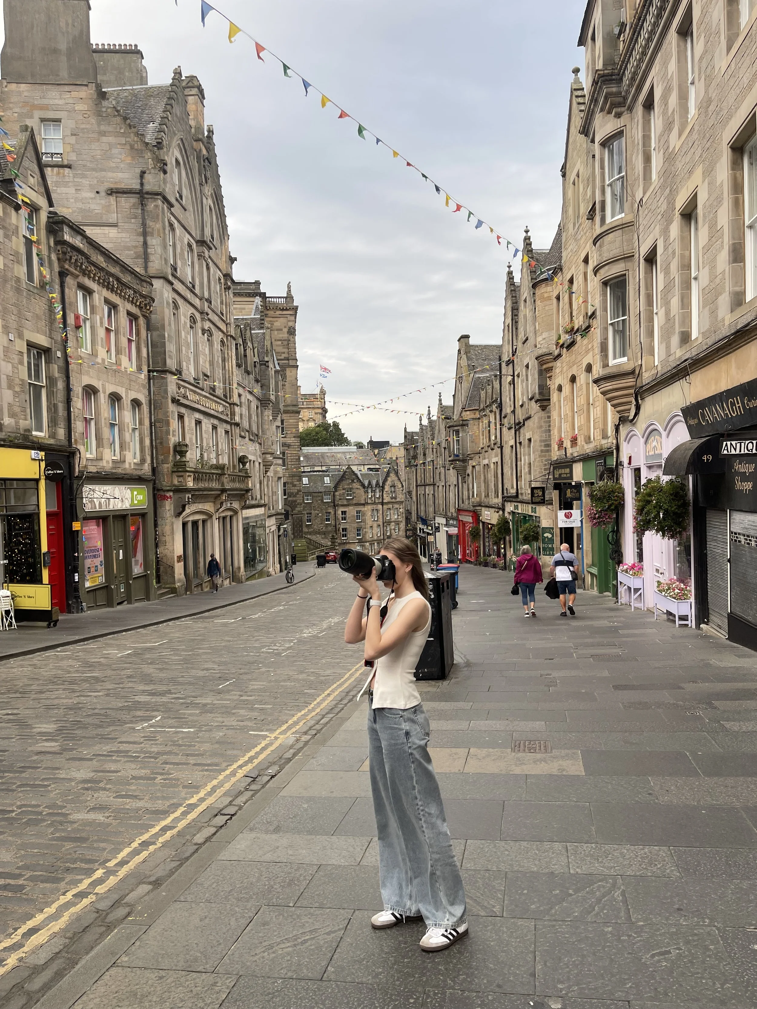 A person standing on a city street taking a photograph with a camera. The street is lined with historic buildings, shops, and colorful bunting decorations hanging overhead. Some pedestrians are walking along the sidewalk.