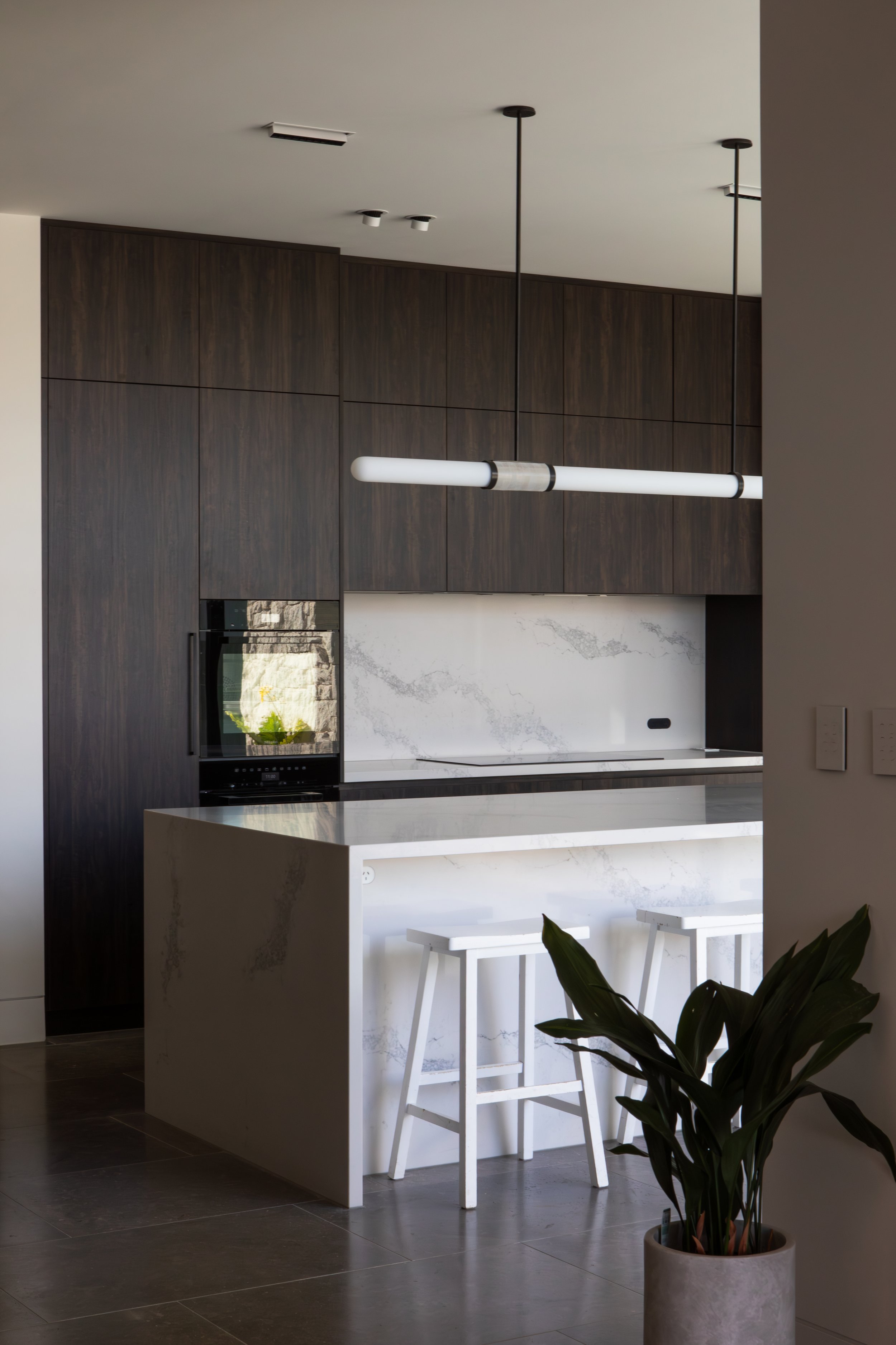 Modern kitchen with dark wood cabinets, a white marble backsplash, a white kitchen island with marble pattern, two white bar stools, a pendant light fixture, and a potted plant in the foreground.