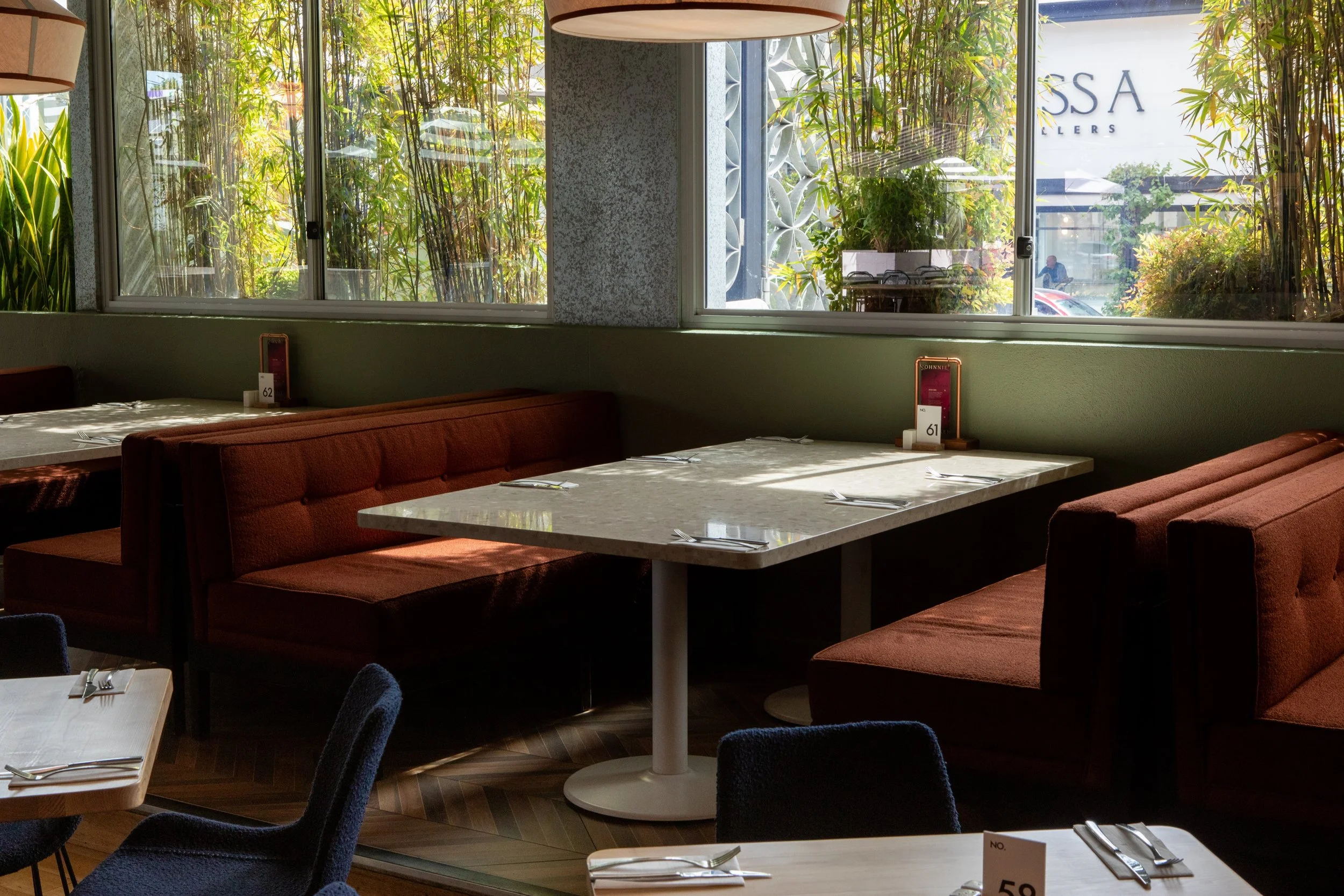 Empty restaurant indoor seating area with brown booths and white marble tables, sunlight coming through large windows with green plants outside.