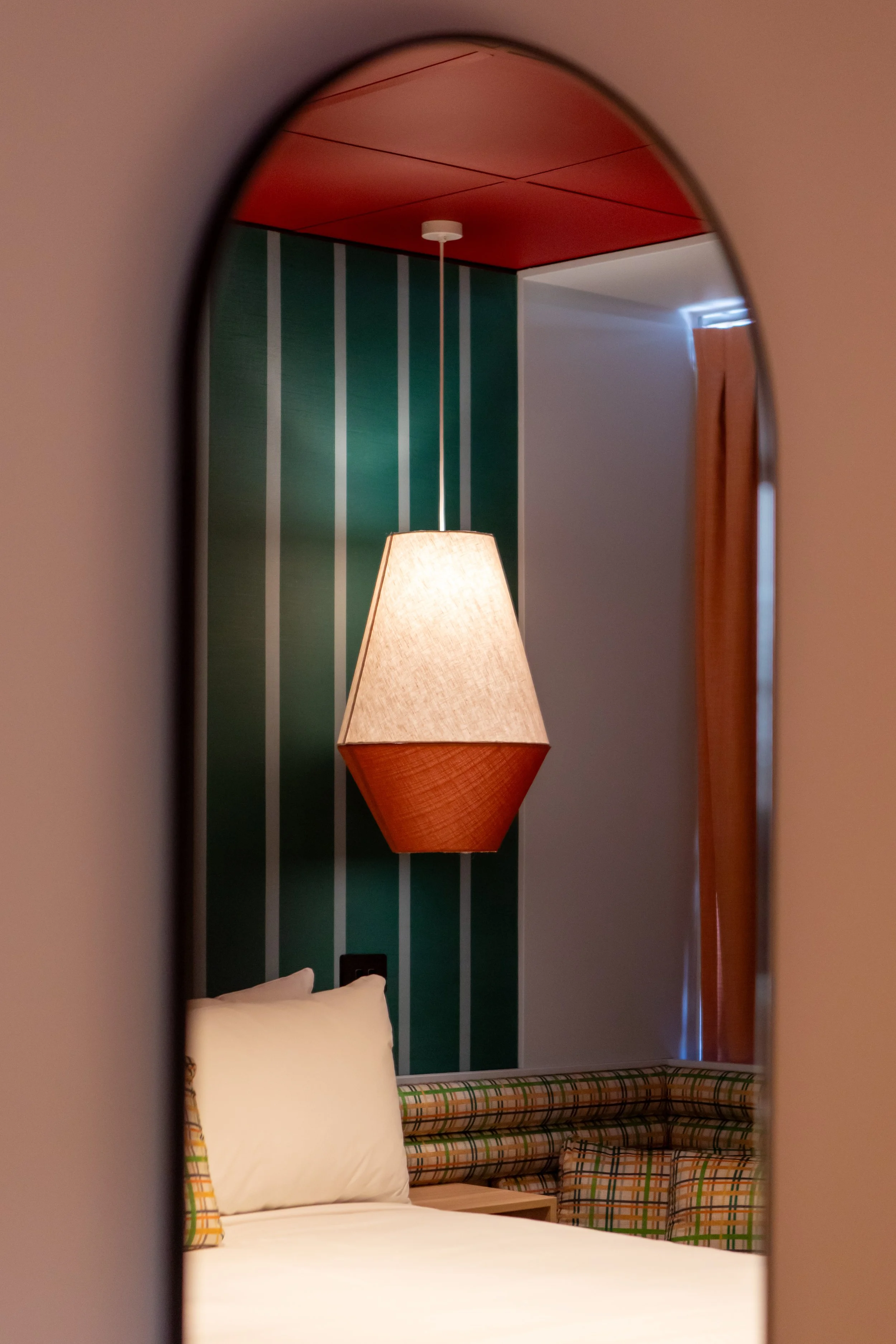 View through an arched mirror showing a bedroom with a hanging lampshade, green striped wall, bed with white bedding, and a corner of patterned furniture.