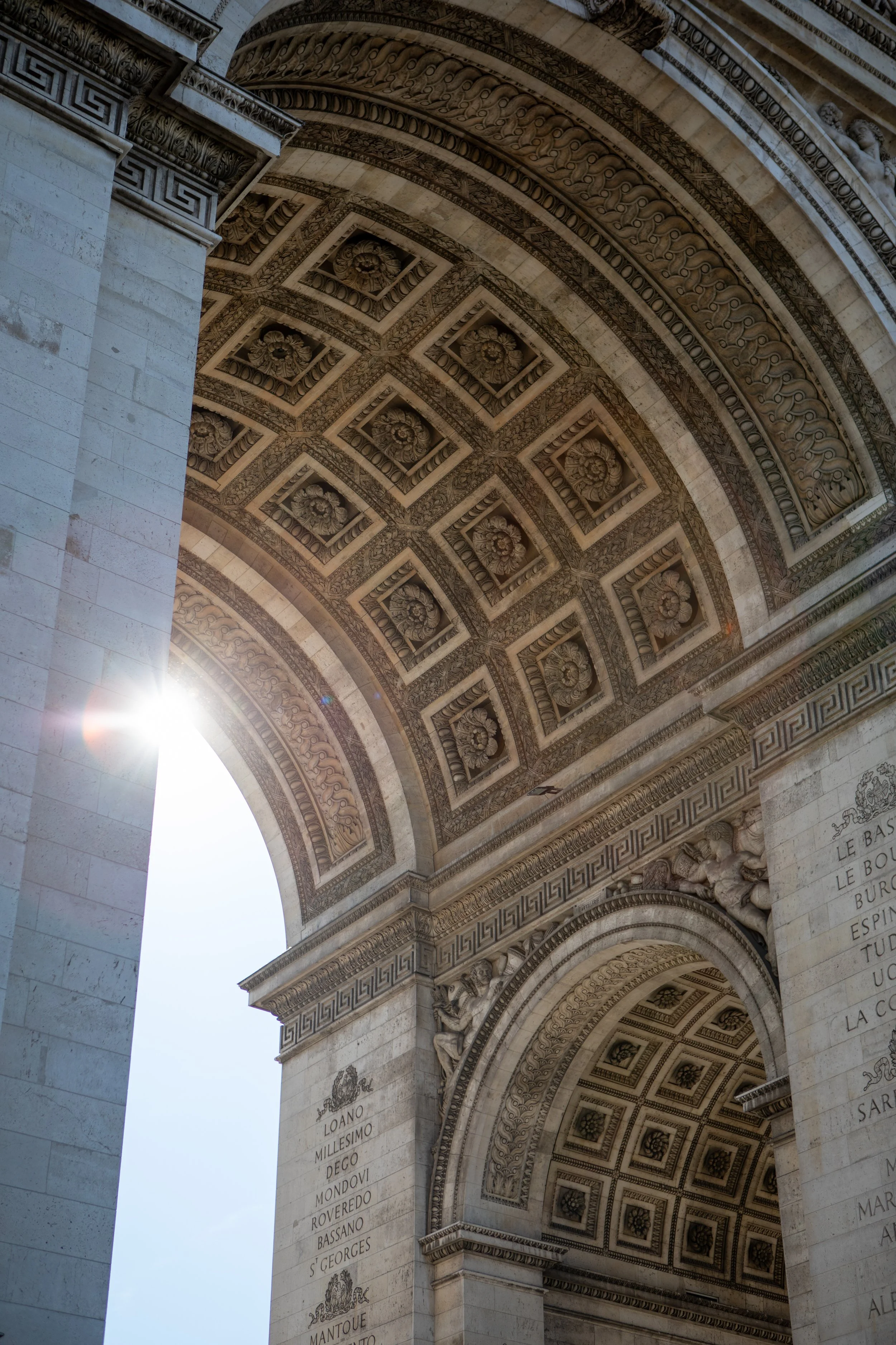 Detailed view of the ornate underside of the Arc de Triomphe, featuring decorative floral patterns and sculpted figures, with sunlight peeking through the upper left corner.