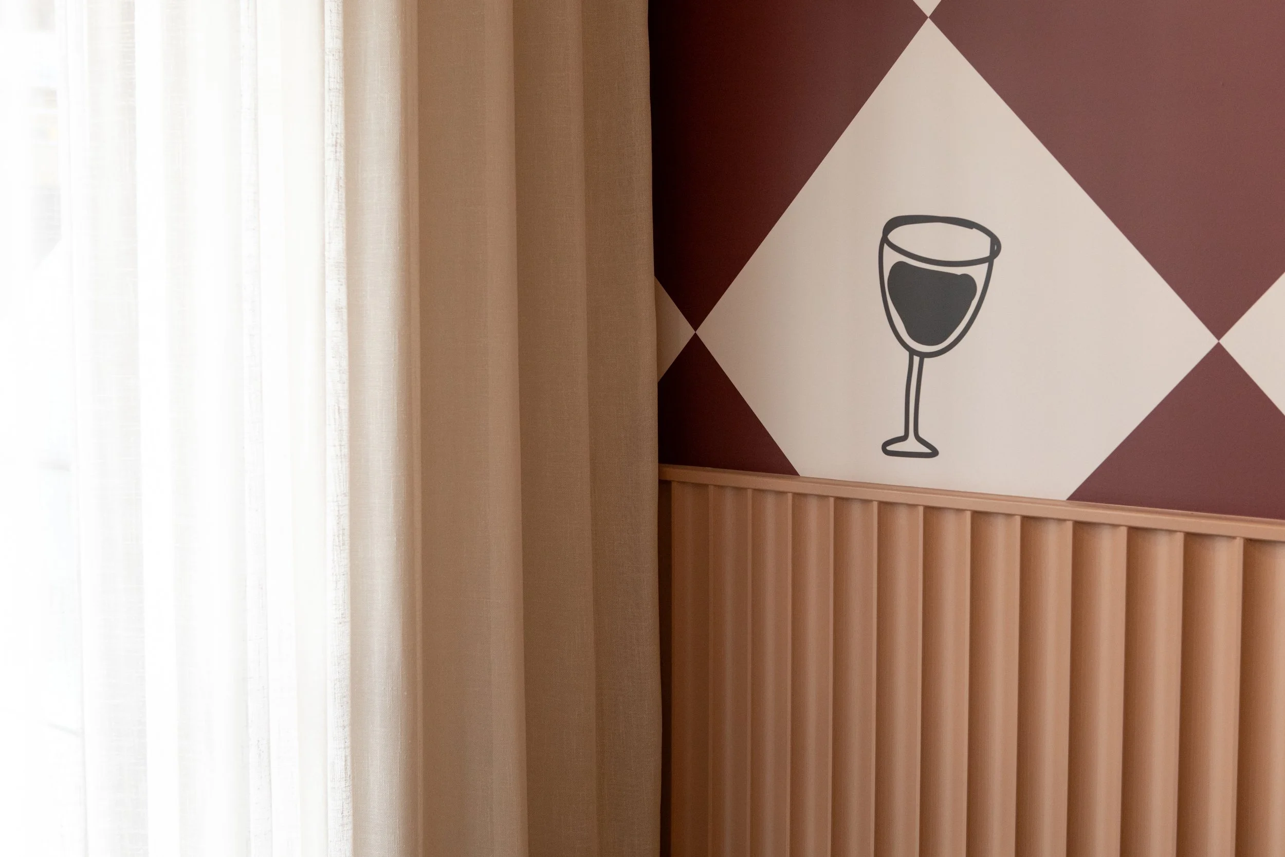 Interior scene with beige curtains on the left, a wall with a decorative pattern of a wine glass on the right, and wooden paneling along the bottom of the wall.