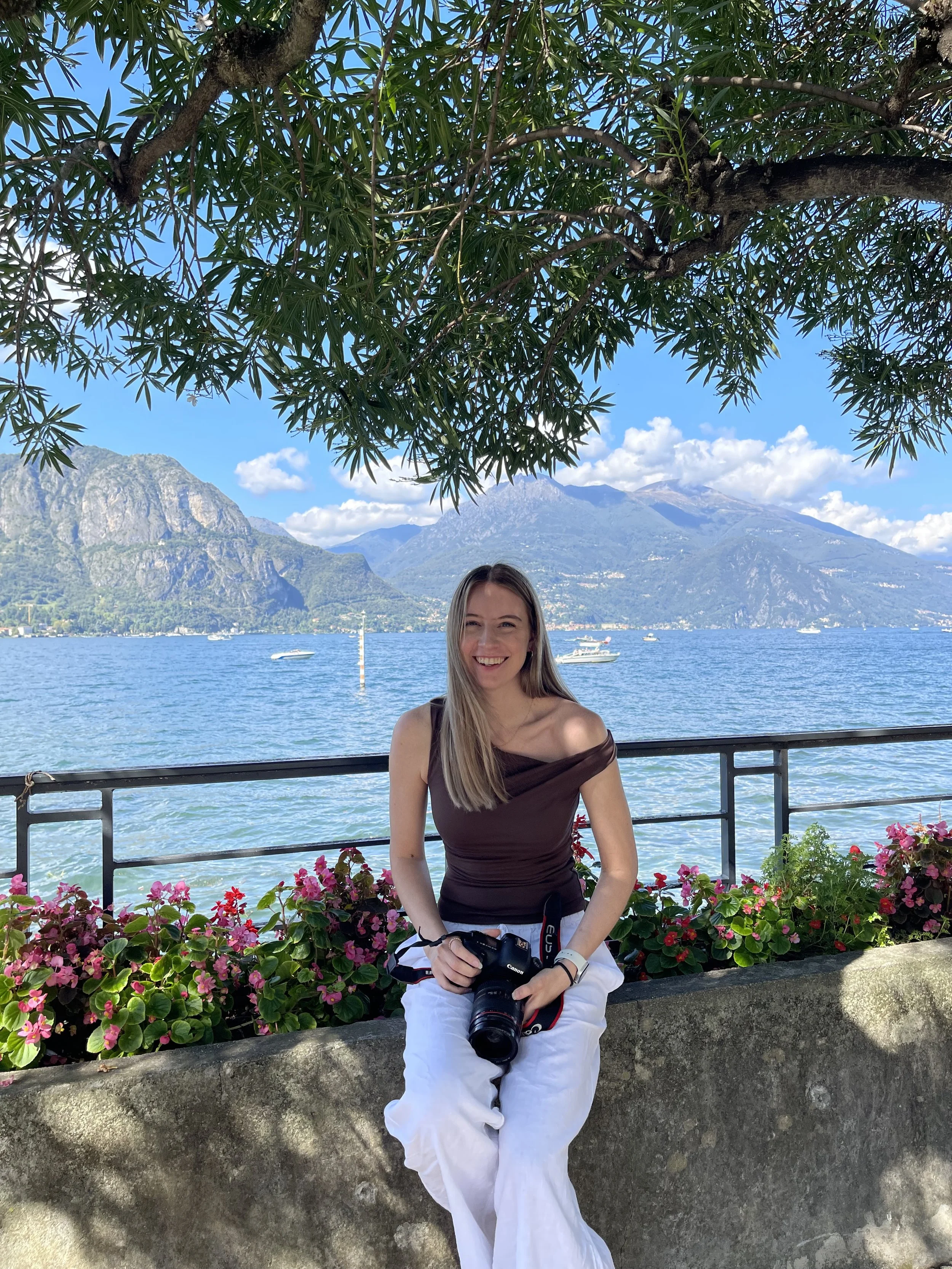 A young woman sits on a stone ledge holding a camera, smiling. She is overlooking Lake Como with mountains in the background, surrounded by pink flowers and green foliage, under a blue sky with some clouds.