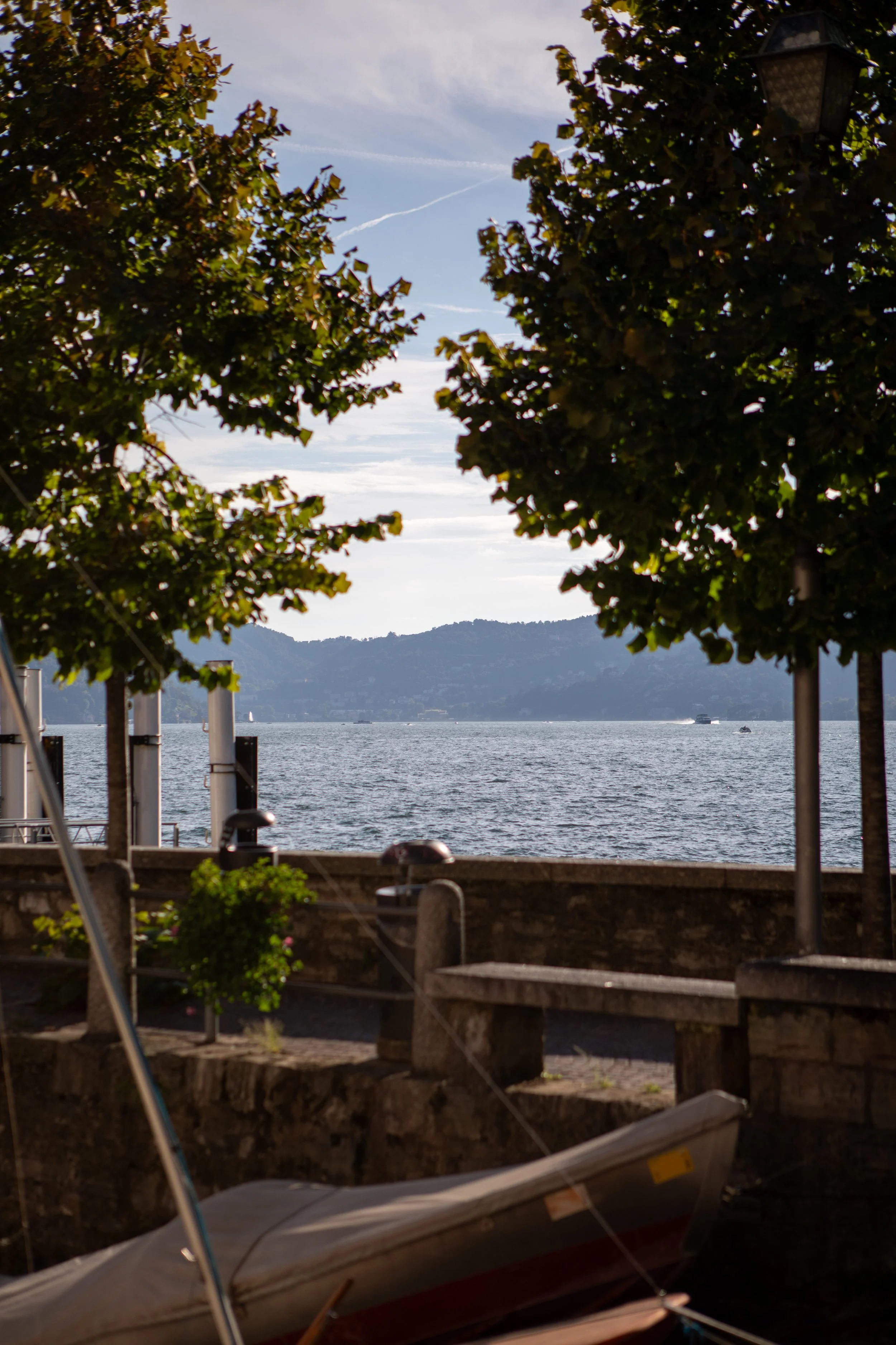 A waterfront scene with a boat in the foreground, trees framing the view, a stone wall with benches and a lamppost, and a body of water with distant hills and a few boats.