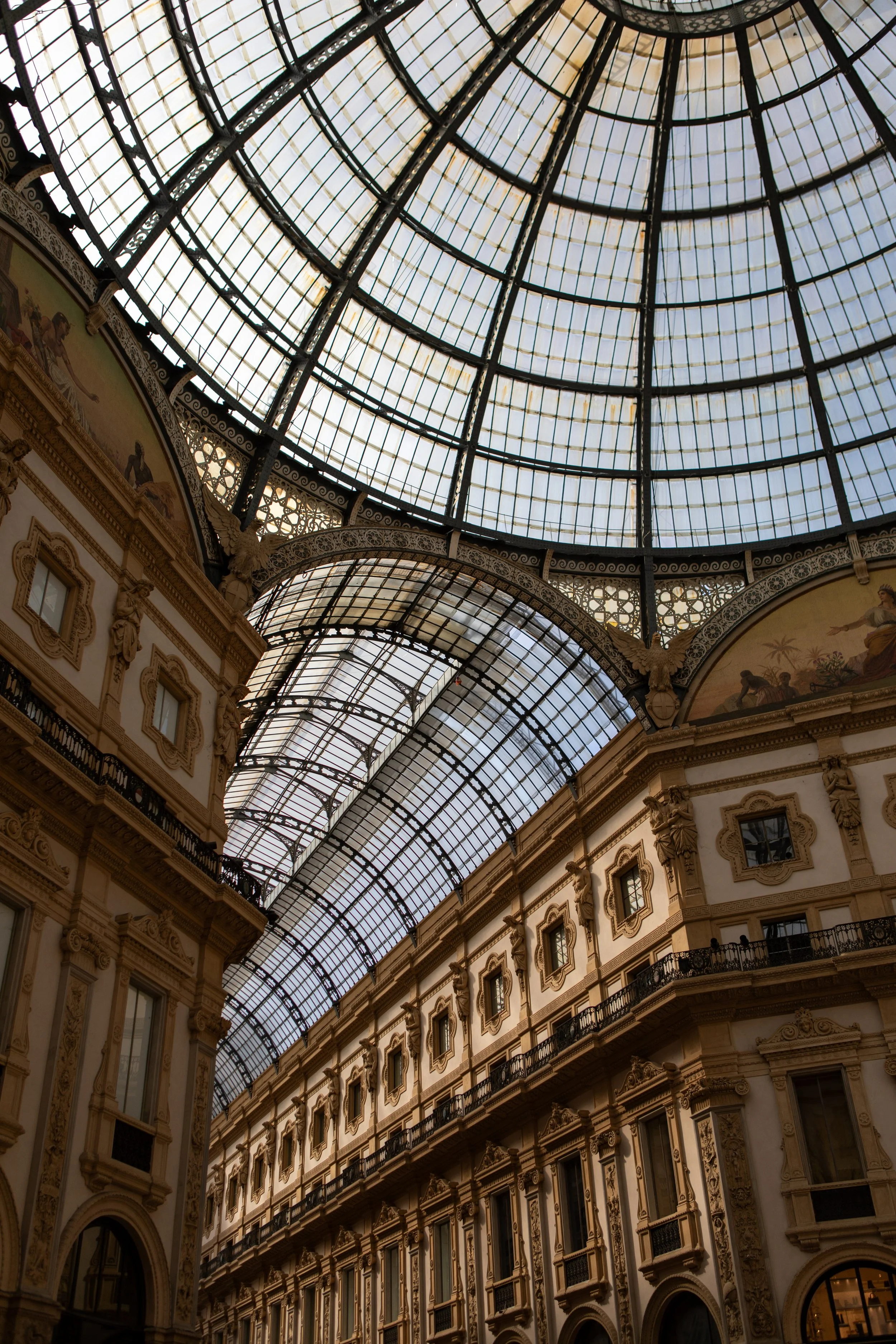 Interior of a grand shopping gallery in Milan with ornate architecture, featuring a large glass dome ceiling and detailed decorative elements on the walls.