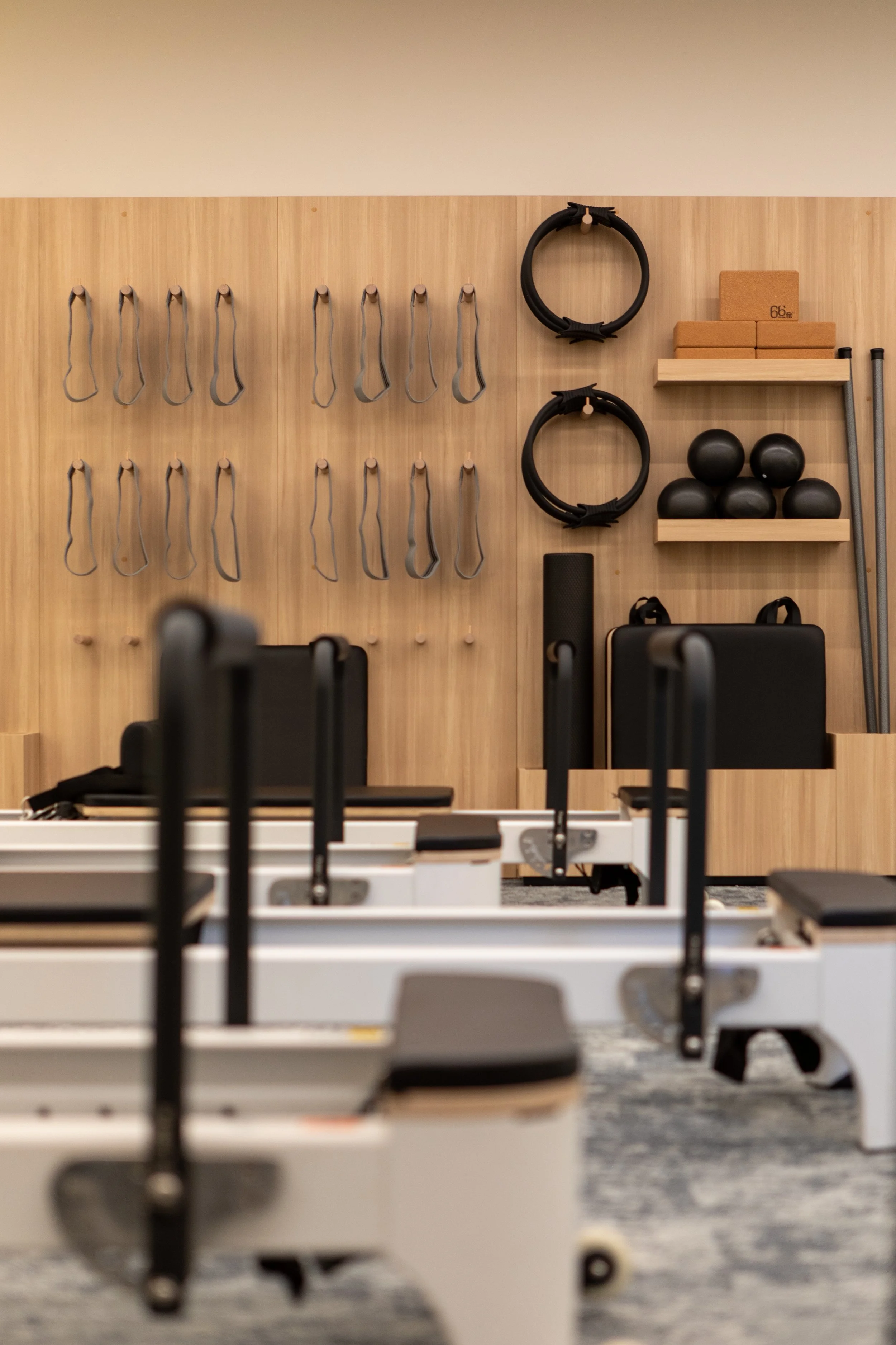 Pilates studio with reformer machines in the foreground and wall-mounted fitness equipment including resistance bands, Pilates rings, yoga blocks, medicine balls, and props in the background.
