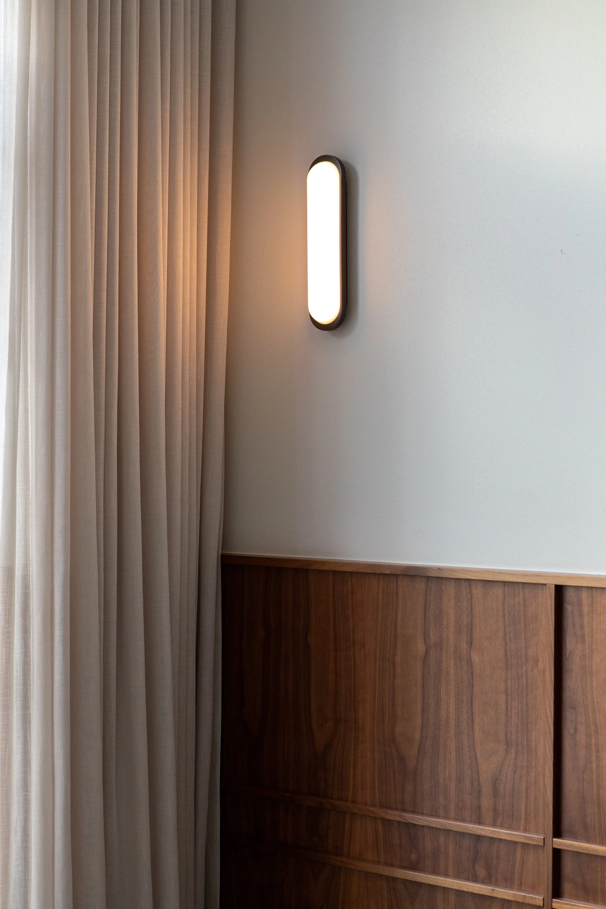 Modern wall-mounted light fixture on a white wall next to beige curtains and wooden paneling.