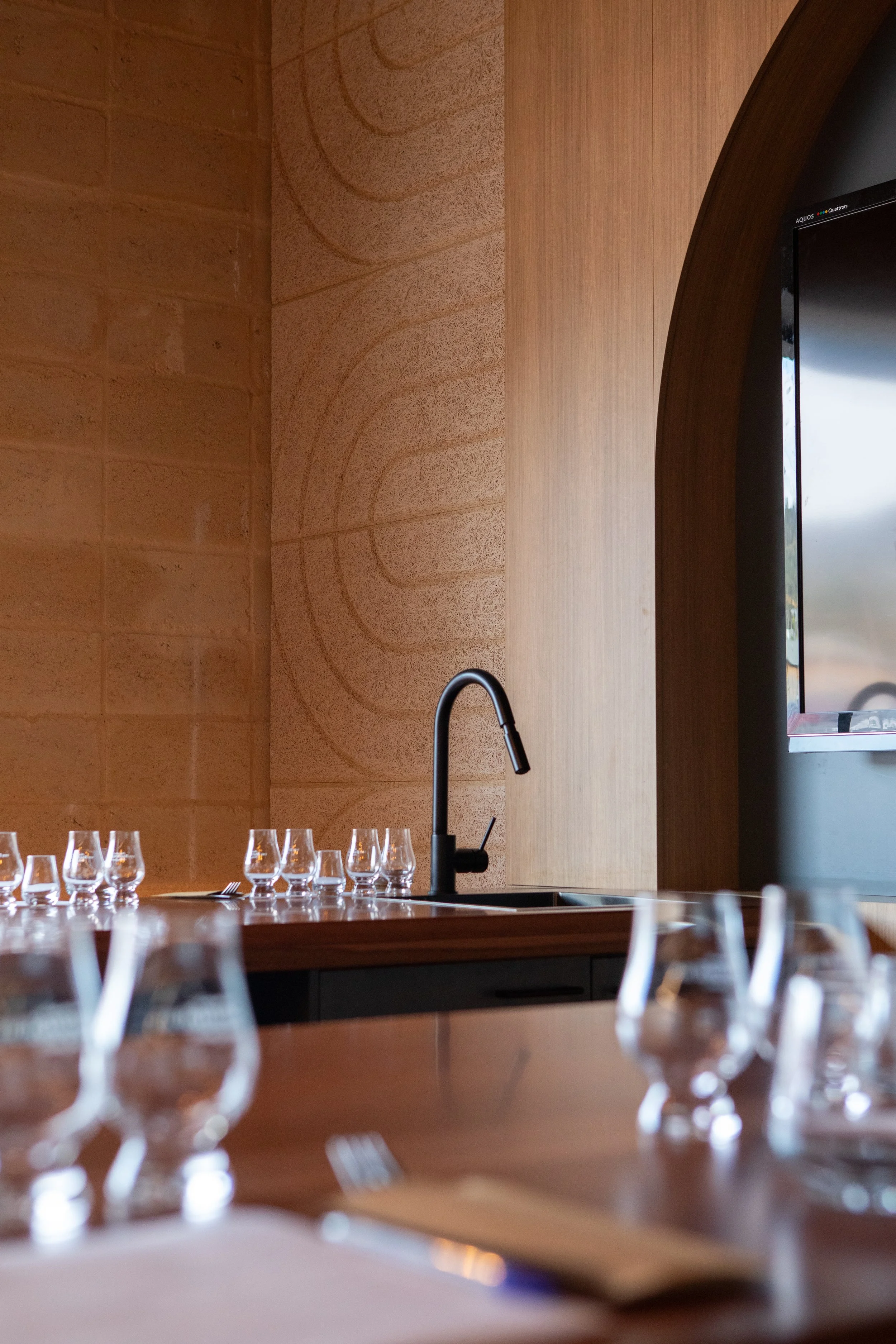 A restaurant or event setup with a wooden table, wine glasses, a water sink, a black faucet, and a wall with textured and patterned surfaces.
