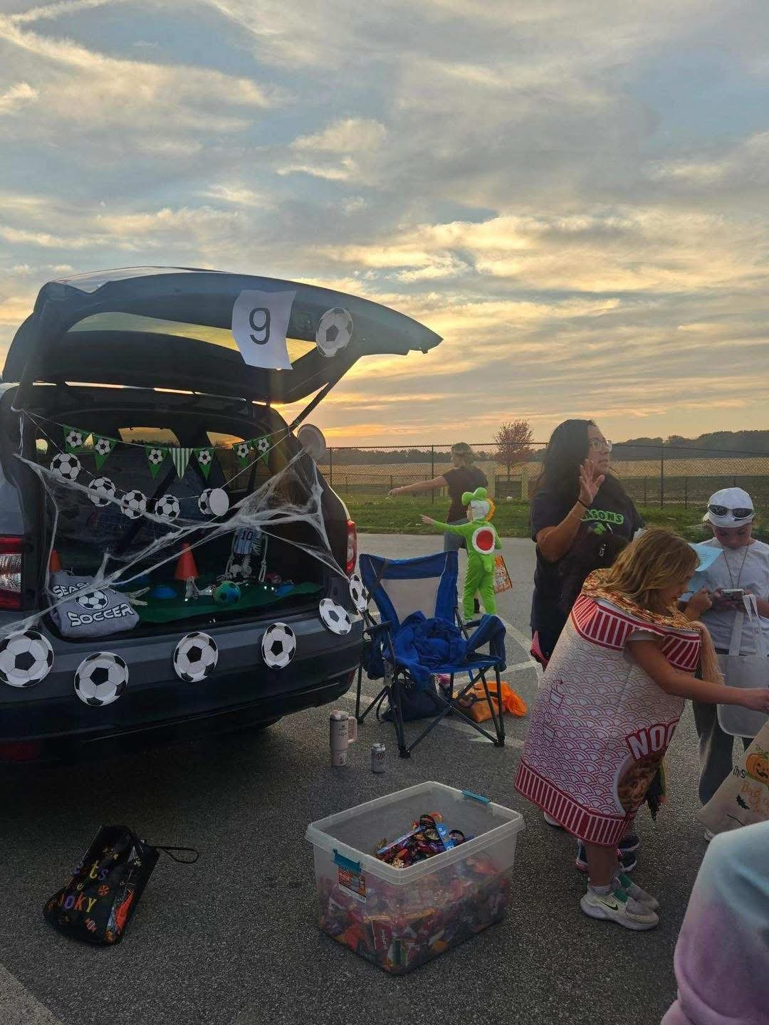 Children and adults at a Halloween-themed outdoor event with a decorated car displaying soccer-themed decorations and costumes, including a person dressed as a frog, during sunset.