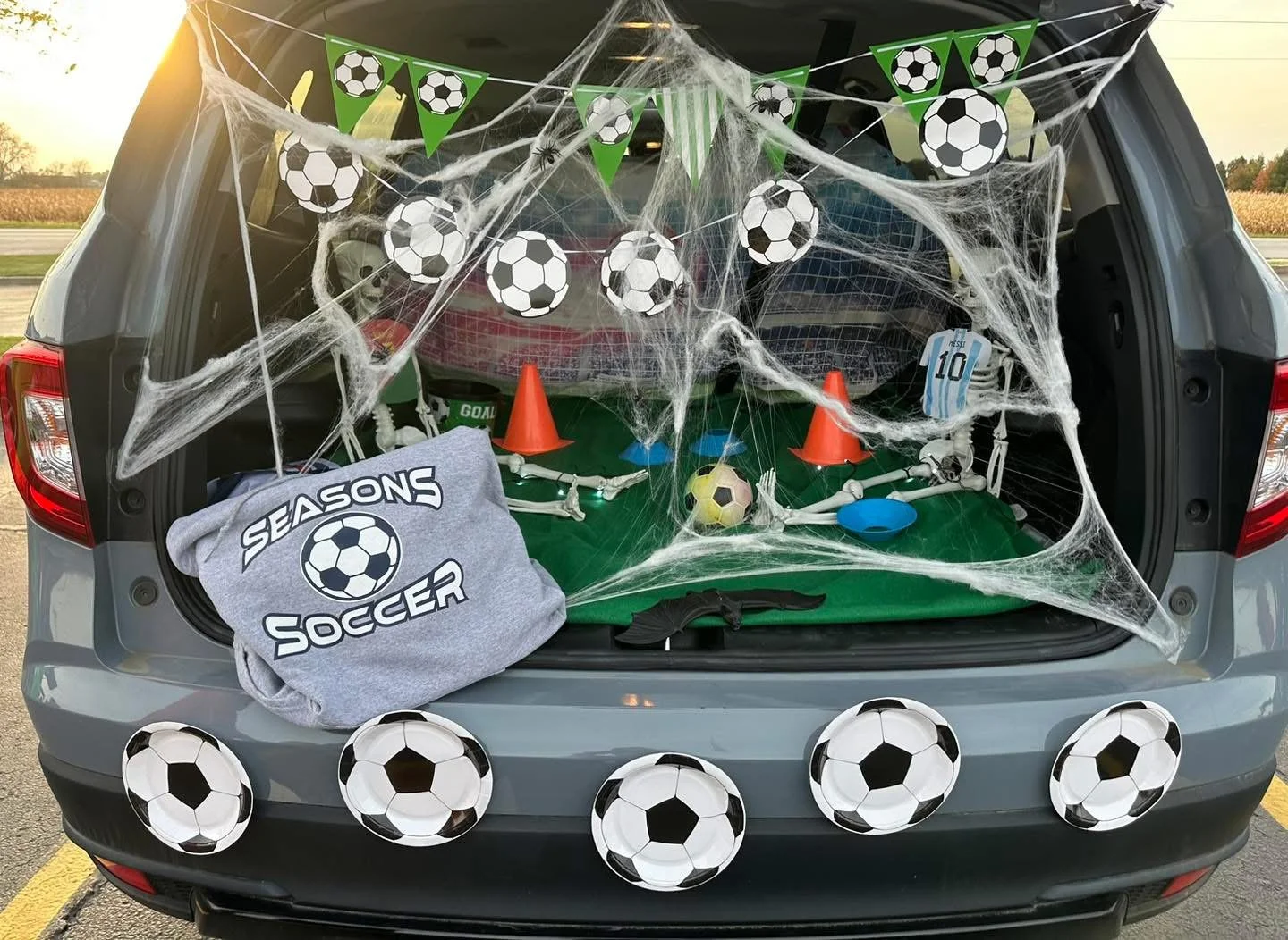 The open trunk of a gray SUV decorated with soccer-themed items including paper soccer balls, orange cones, a small goal, a jersey with the number 10, and a gray sock with a soccer ball and the words "Seasons Soccer". There are cobweb decorations and soccer balls placed around the trunk.