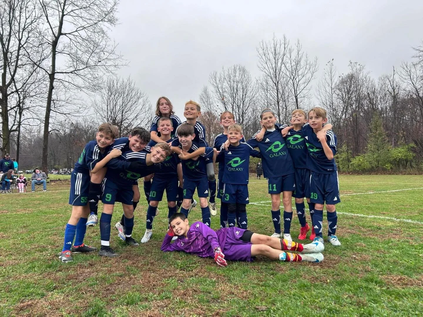 A group of young boys in soccer uniforms pose together on a sports field, some with arms around each other, with a boy in a purple goalkeeper jersey lying in front. They are smiling, and there are trees and spectators in the background.