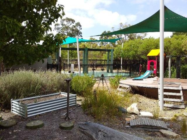 Outdoor playground with slides, shade sails, garden beds, and wooden seating area; surrounded by trees and bushes.
