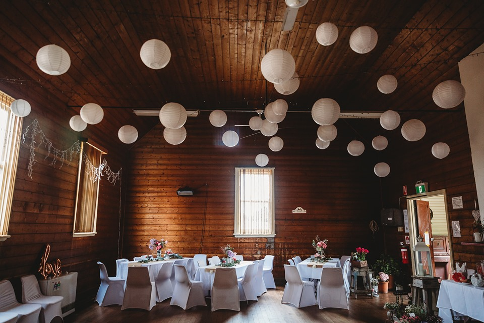Rustic interior with wooden walls and ceiling decorated with white paper lanterns. The room has tables covered with white cloths and chairs with white covers, adorned with floral centerpieces. Natural light enters through windows, enhancing the cozy 
