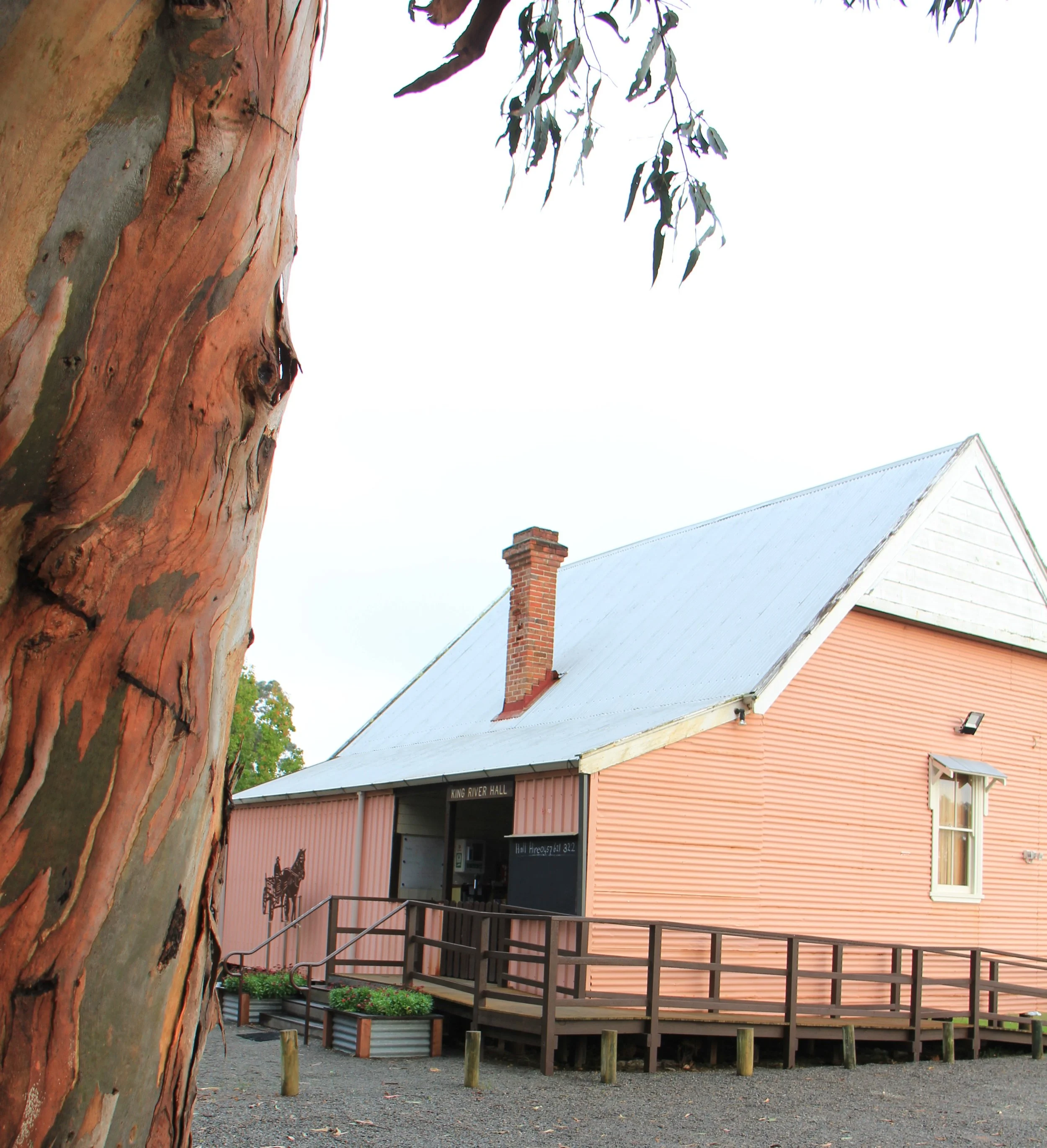 Rustic building with a corrugated metal exterior, labeled "King River Hall," and surrounded by a wooden porch. A large tree trunk is in the foreground.