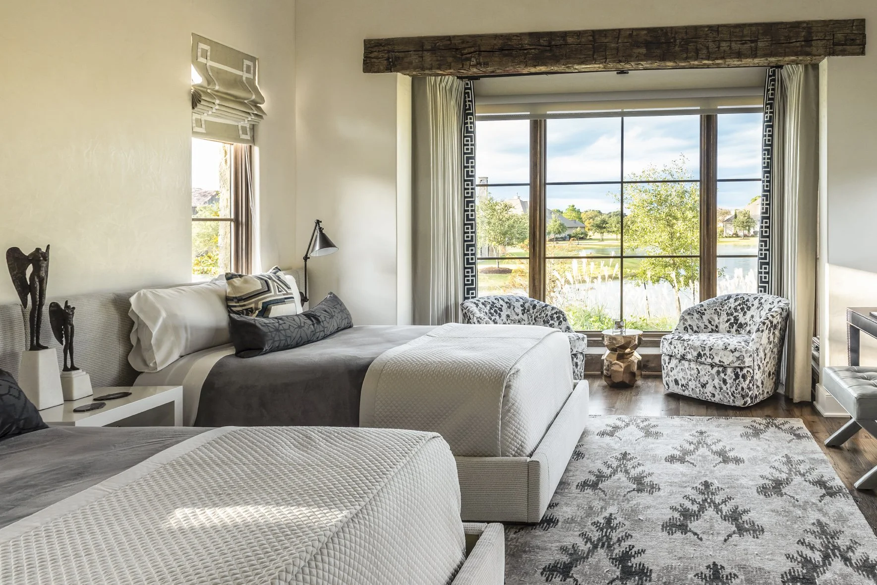 Texas ranch twin bedroom with layered gray bedding, patterned swivel chairs, dark wood floors and a lake-facing window.
